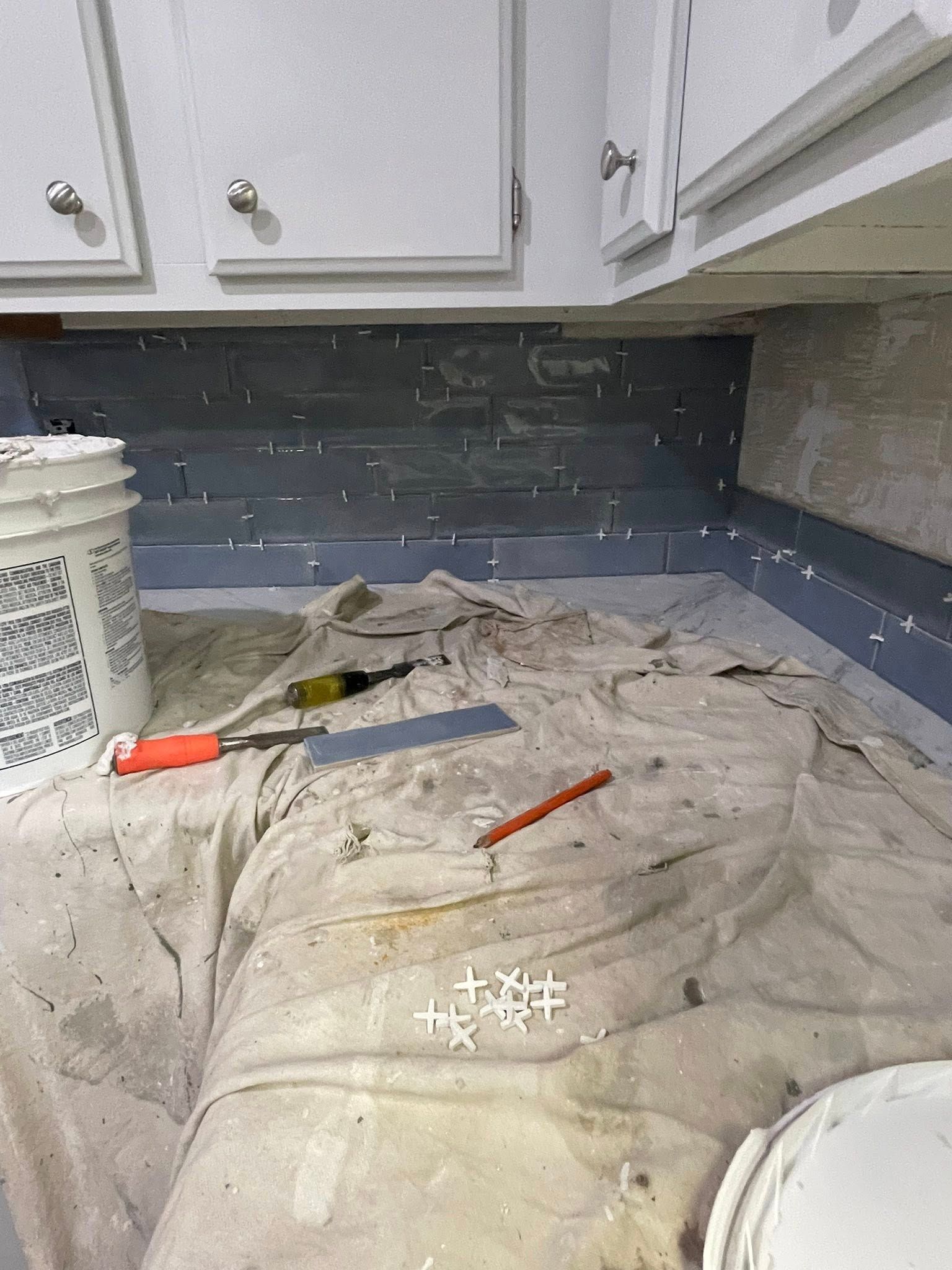 Tile installation in progress under white kitchen cabinets; gray tiles with spacers, tools, and bucket.