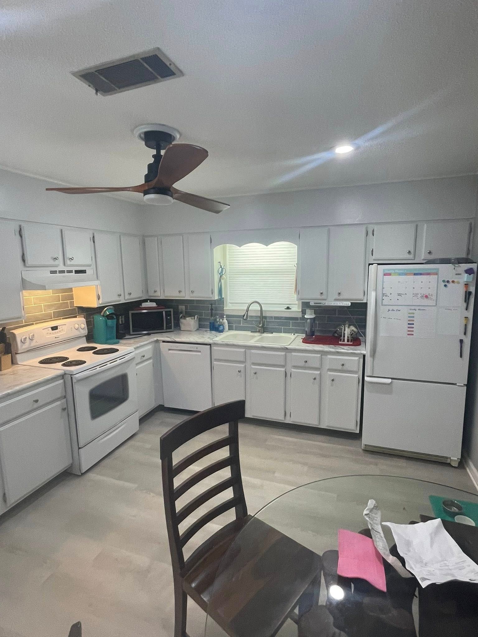 White kitchen with white appliances, cabinets, and a ceiling fan. A dark wooden chair sits at a glass table.
