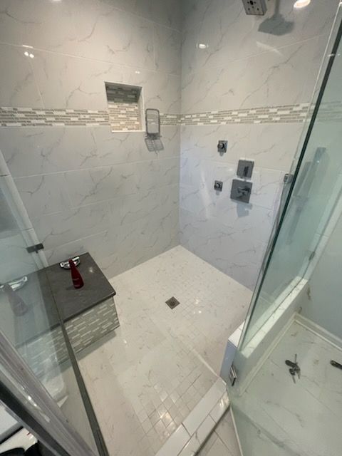 Spacious tiled shower with glass door and bench. White and gray tones, modern design.