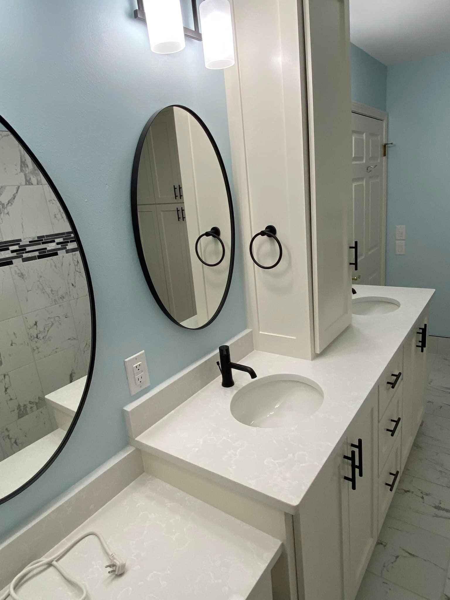 Bathroom with light blue walls, white cabinets, oval mirrors, and black fixtures.