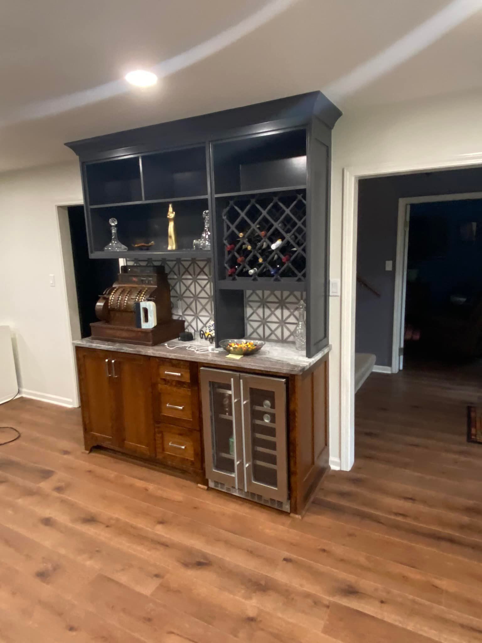 Built-in home bar with wood cabinets, wine storage, and a mini-fridge. Dark gray shelving above, marble countertop, and patterned backsplash.