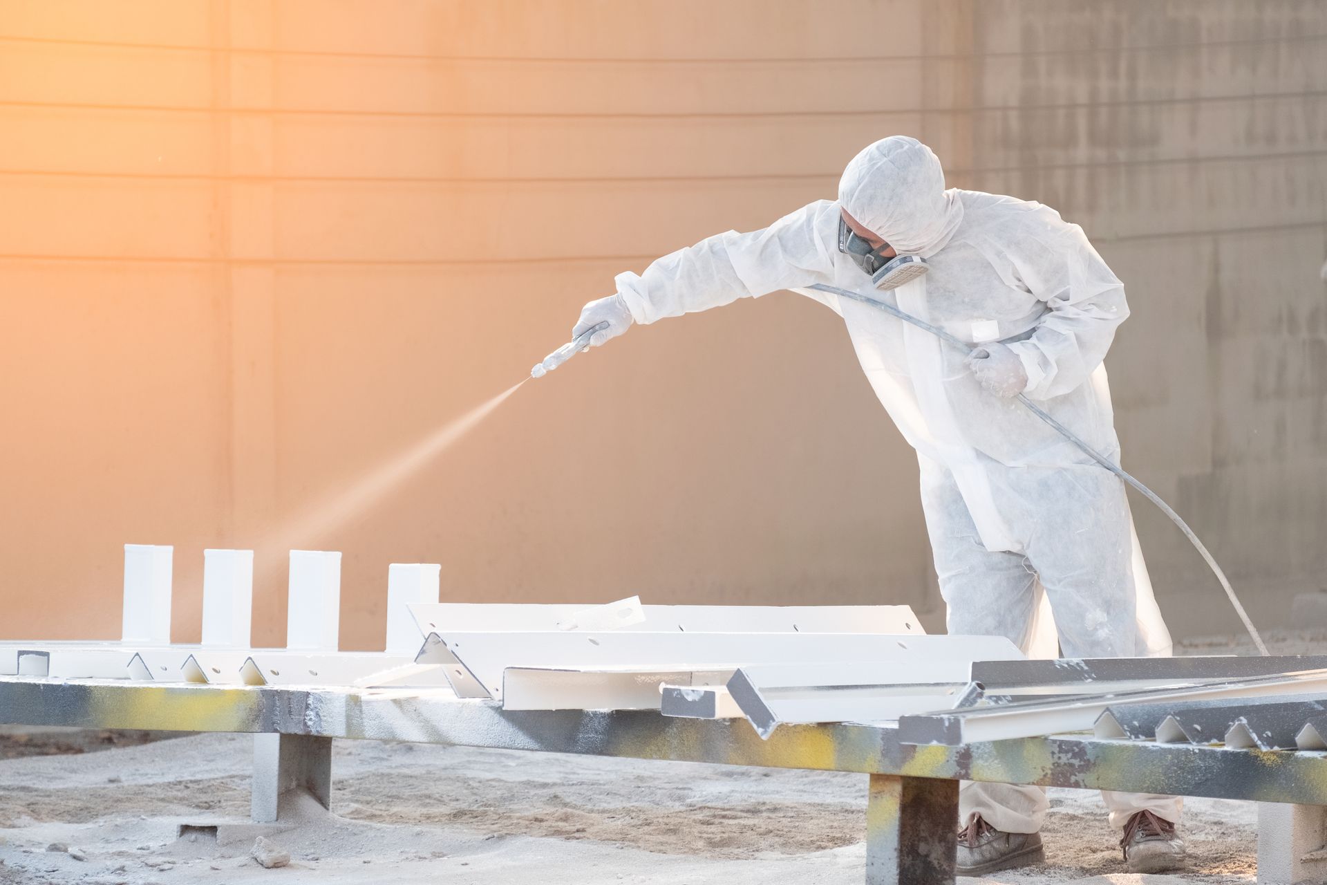 Person in white protective suit spraying white paint on metal beams outdoors.