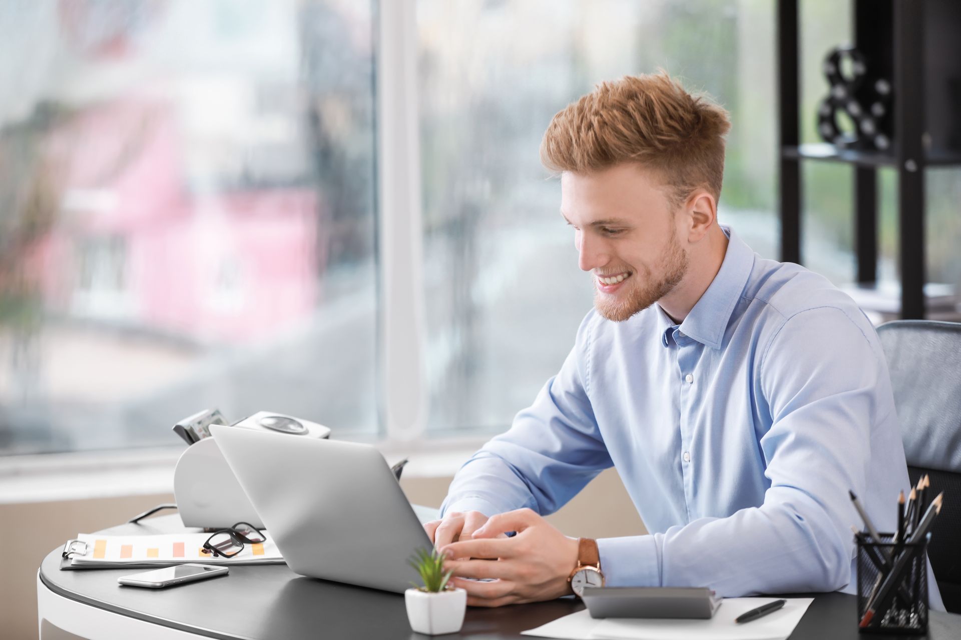 Man smiling while working on a laptop at a desk near a window.