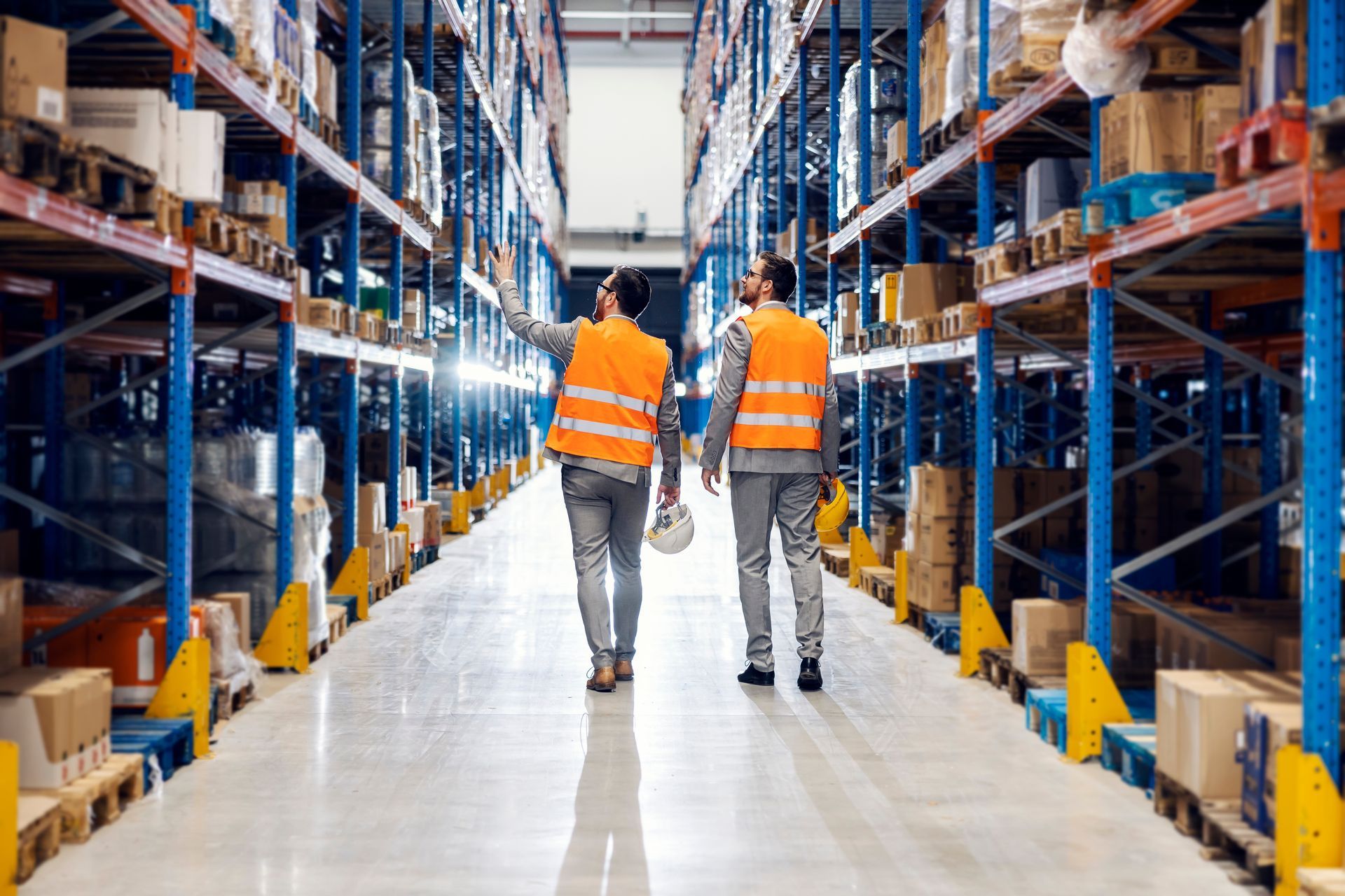 Two workers in orange vests walk down a warehouse aisle lined with shelves of inventory.