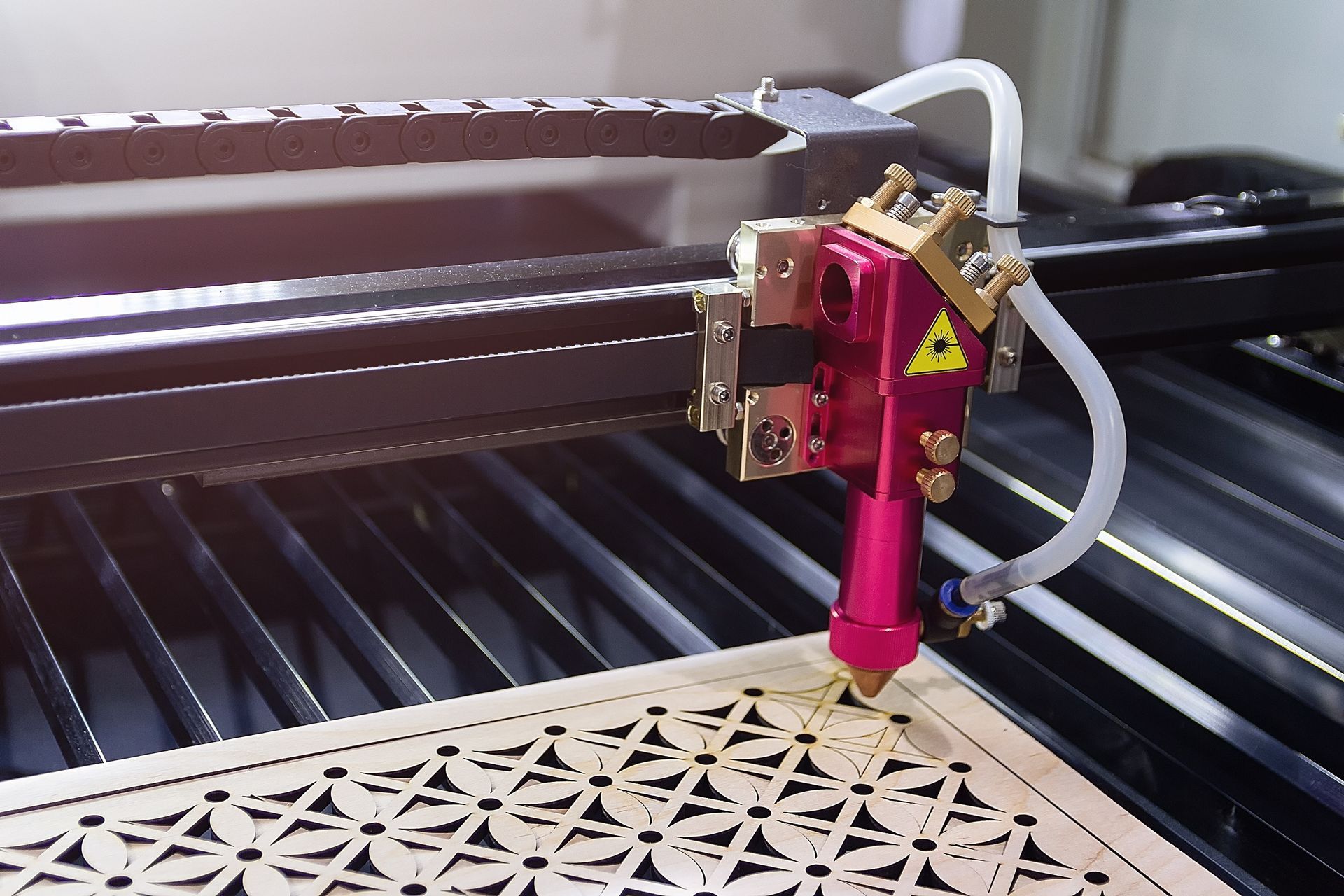 Laser cutter cutting a geometric pattern in wood. Pink laser head, black machine, close-up.