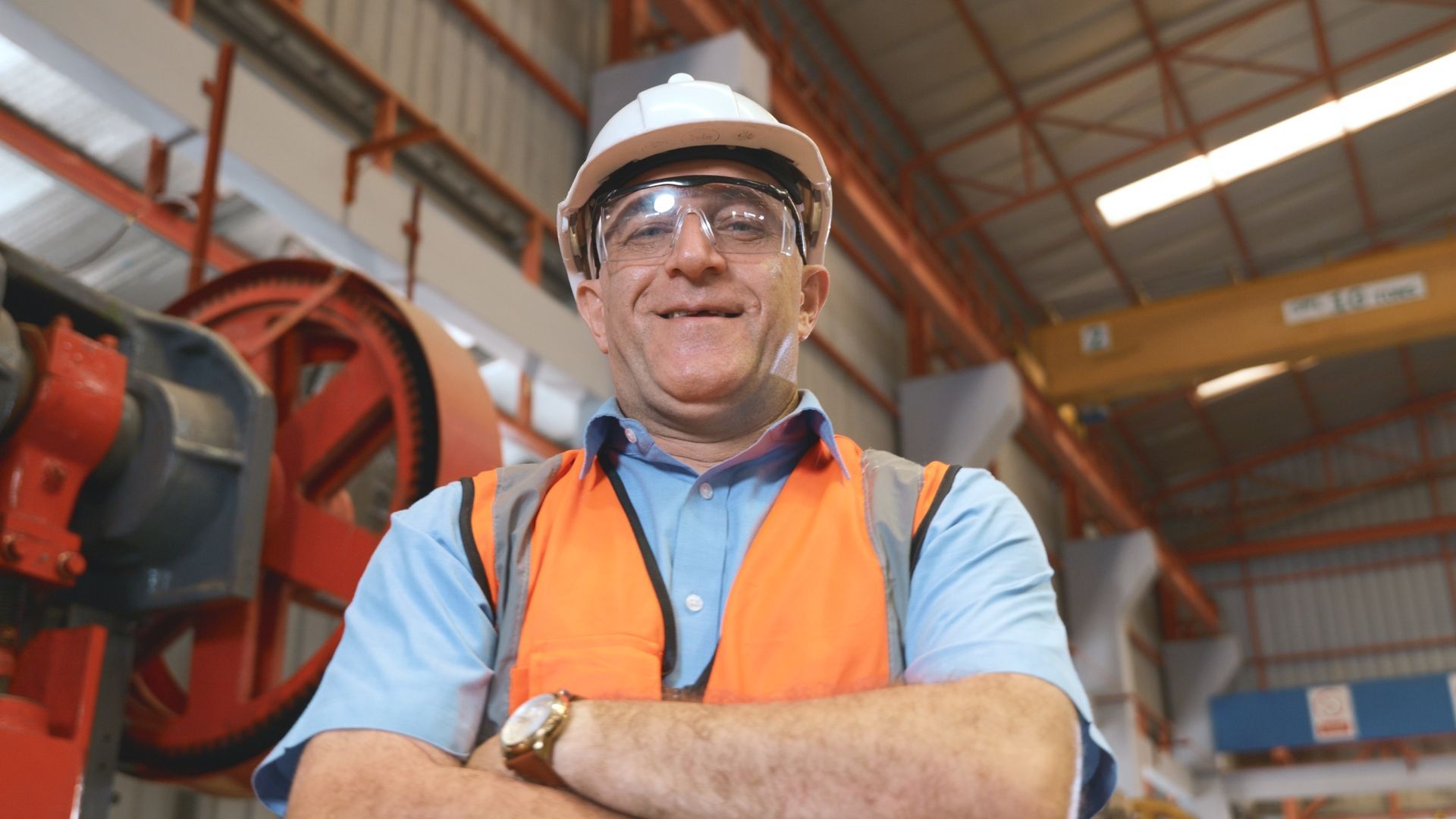 Man in safety gear, arms crossed, smiles. Inside industrial building with machinery.