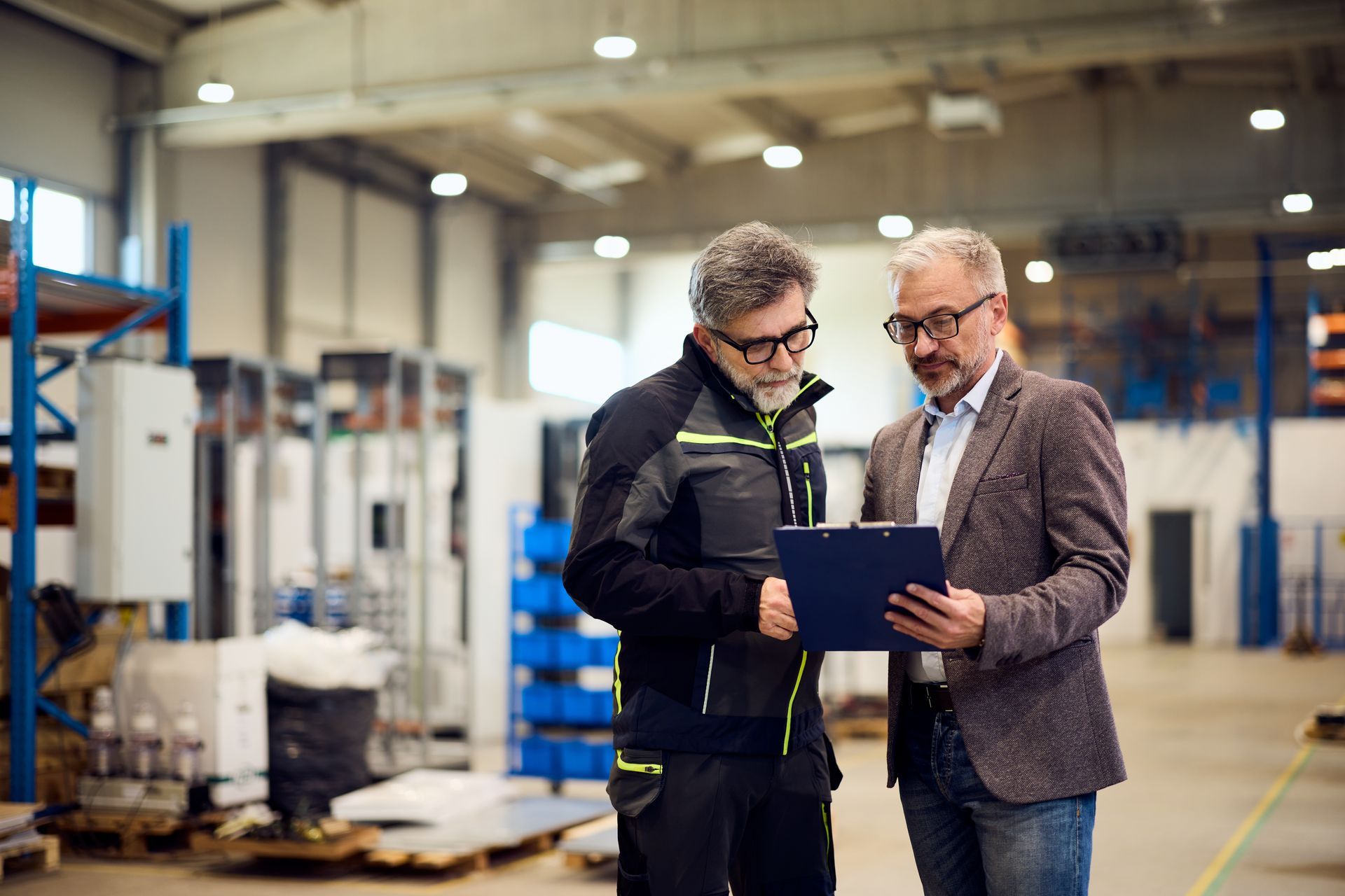 Two men in a warehouse reviewing documents on a clipboard. One wears work attire, the other a blazer.