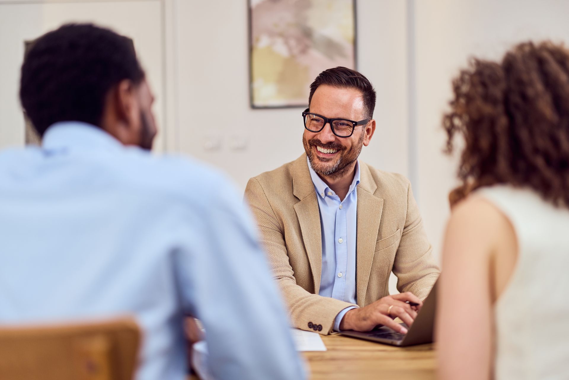 Smiling man in glasses at desk with laptop, talking to two people.