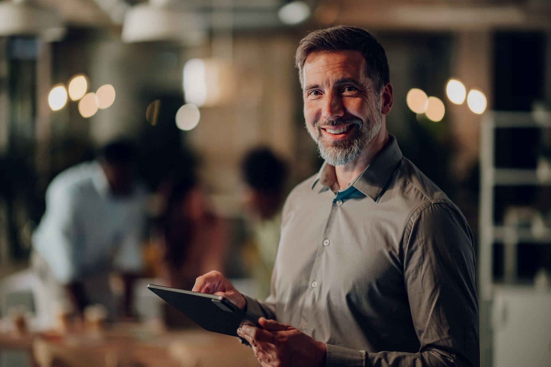 Man in button-up shirt smiling while holding a tablet in an office setting with people blurred in the background.