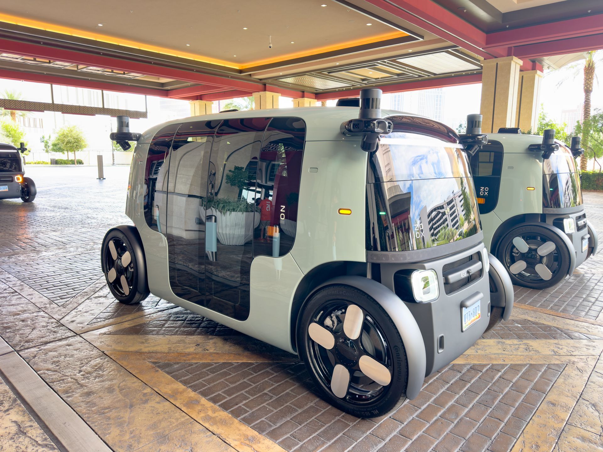 Two autonomous, olive-green passenger vehicles parked under a portico. They have large windows and no driver's seat visible.