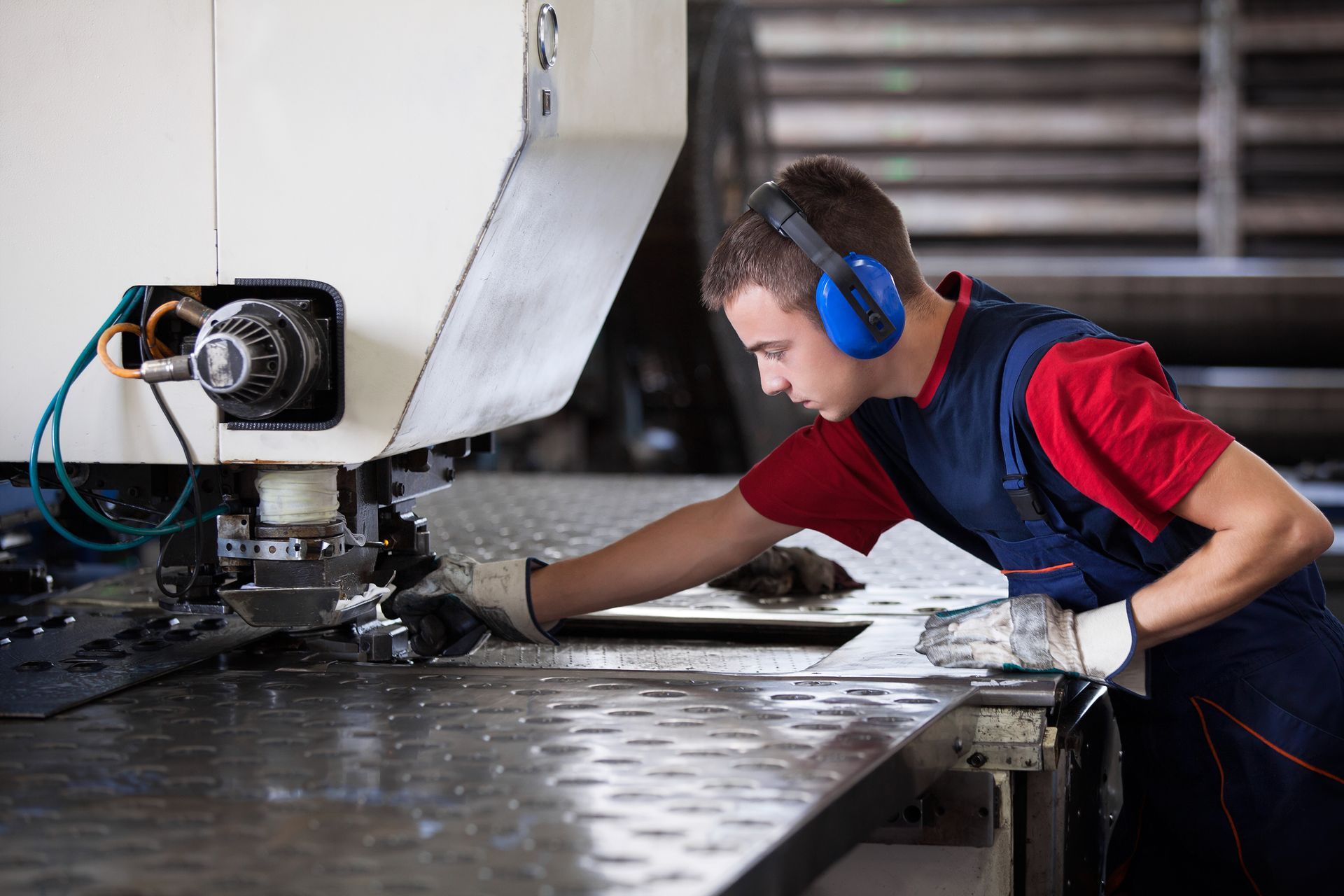 Worker operating industrial punch press, wearing ear protection, and protective gloves in a factory.
