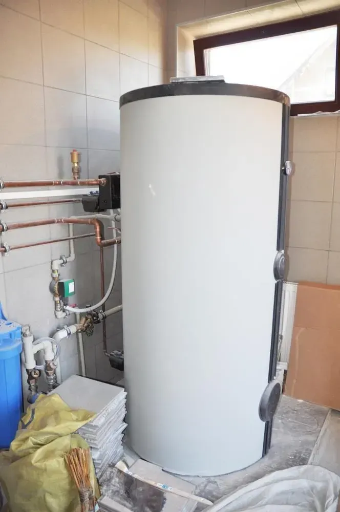 Large white cylindrical water heater in a tiled utility room with pipes and a blue barrel