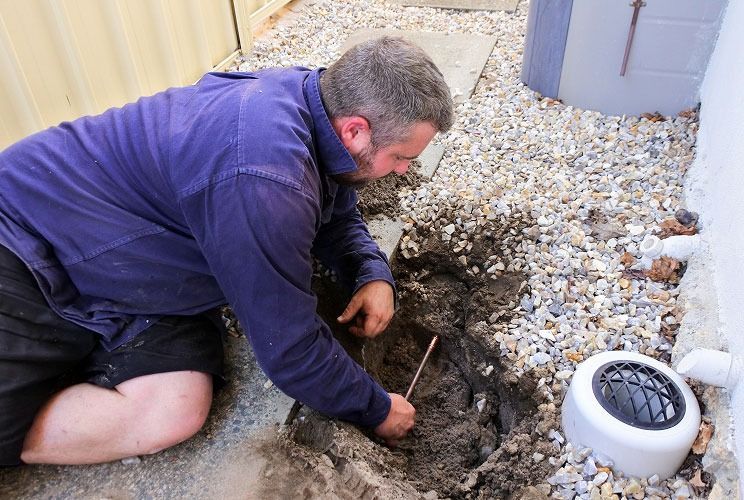 Man kneeling in a gravel yard, digging into a hole beside a white vent pipe