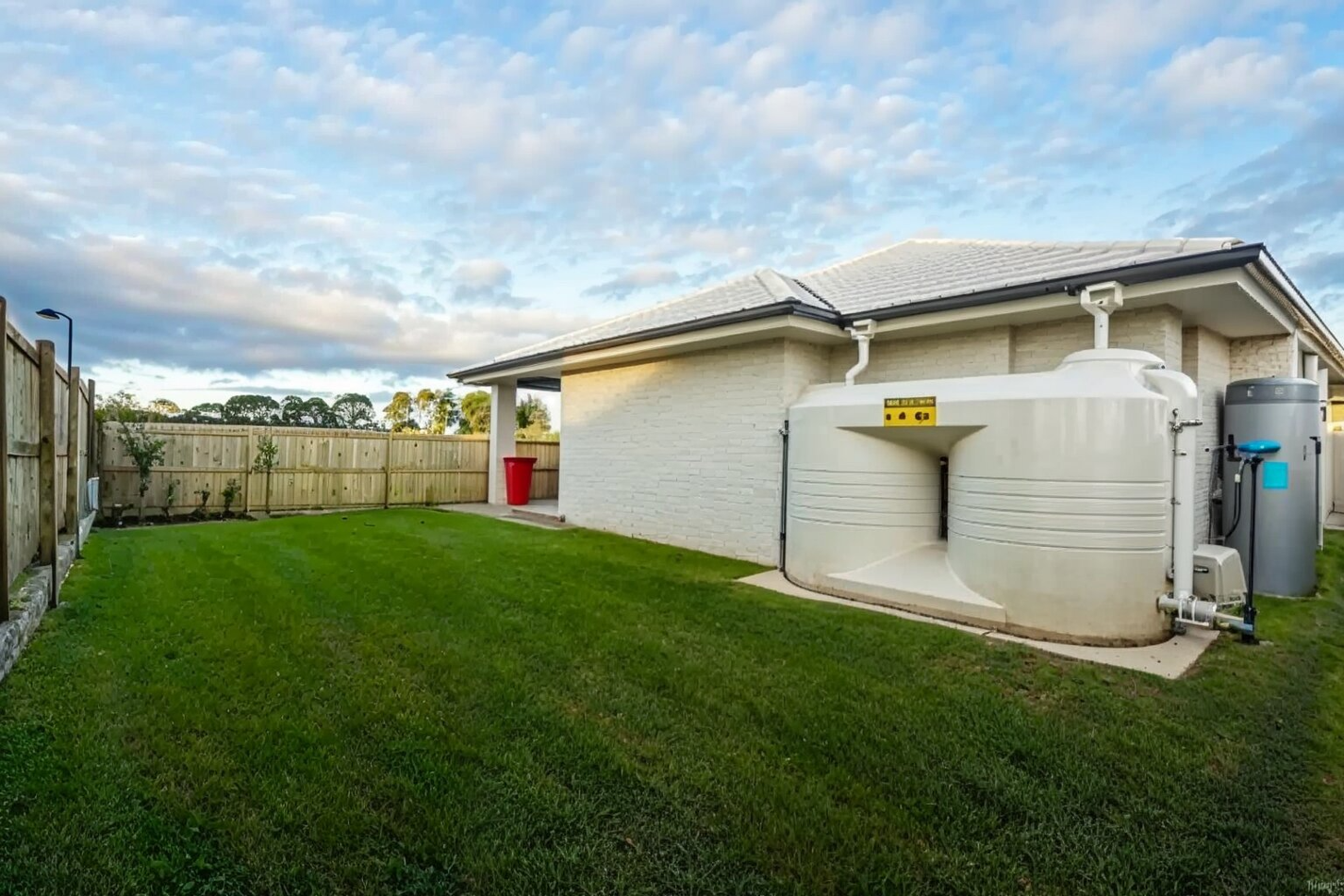 Backyard with green lawn, white house, wooden fence, and large water tanks by the wall.