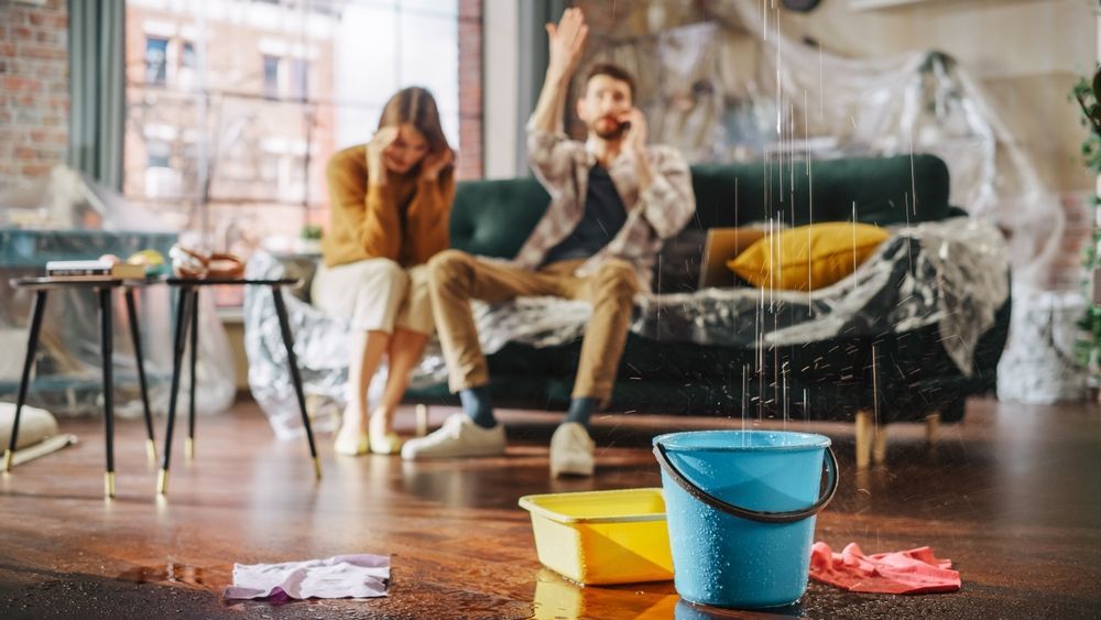 Buckets and spilled water on a hardwood floor, with people sitting on a couch in the background inside a flooded room