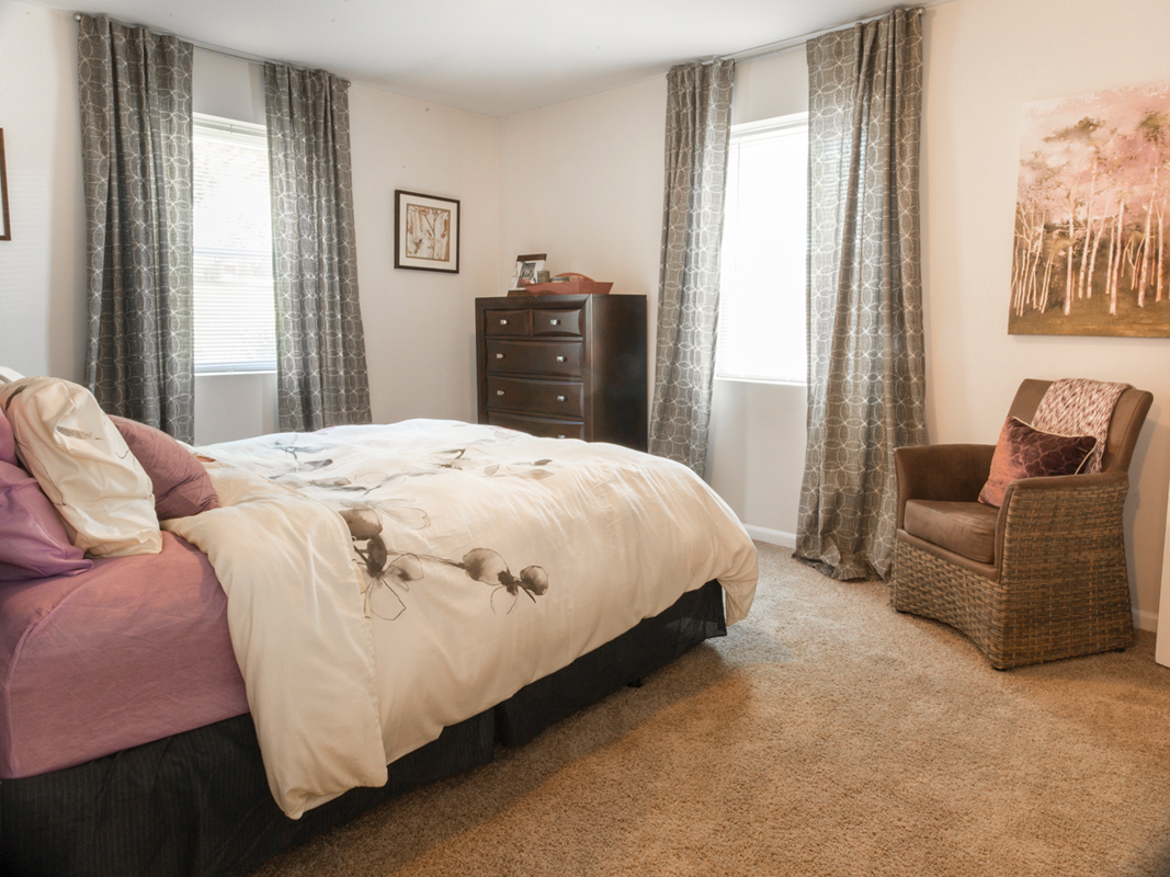 A bedroom with a bed, dresser, chair and two windows.