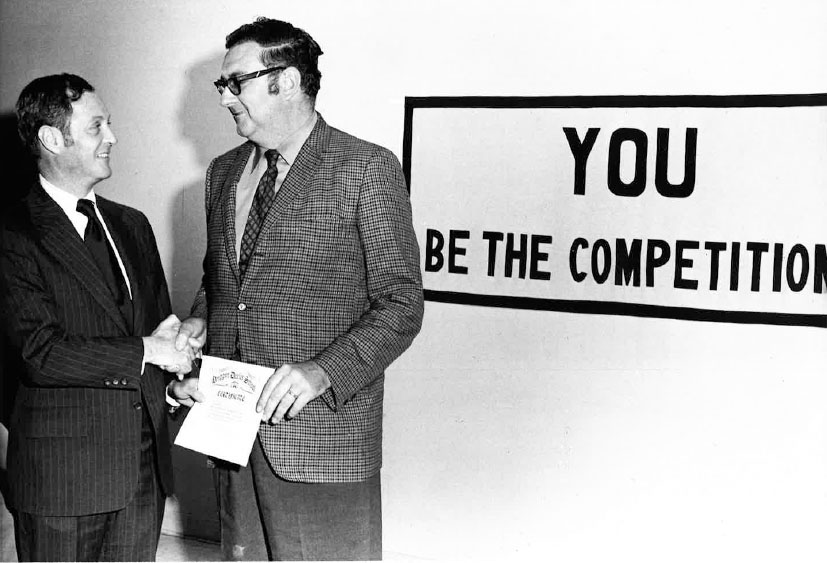 Two men shaking hands in front of a sign that says you be the competition