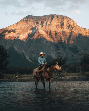 A man is riding a horse in a river with mountains in the background.
