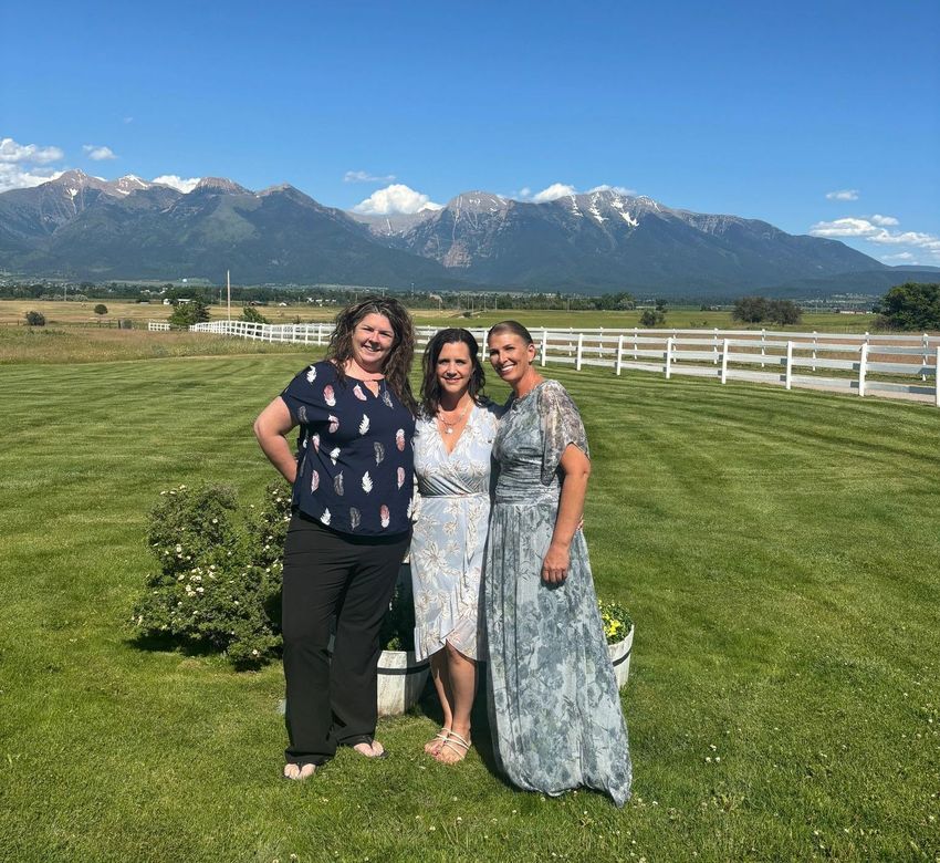 Three women are posing for a picture in a grassy field with mountains in the background.