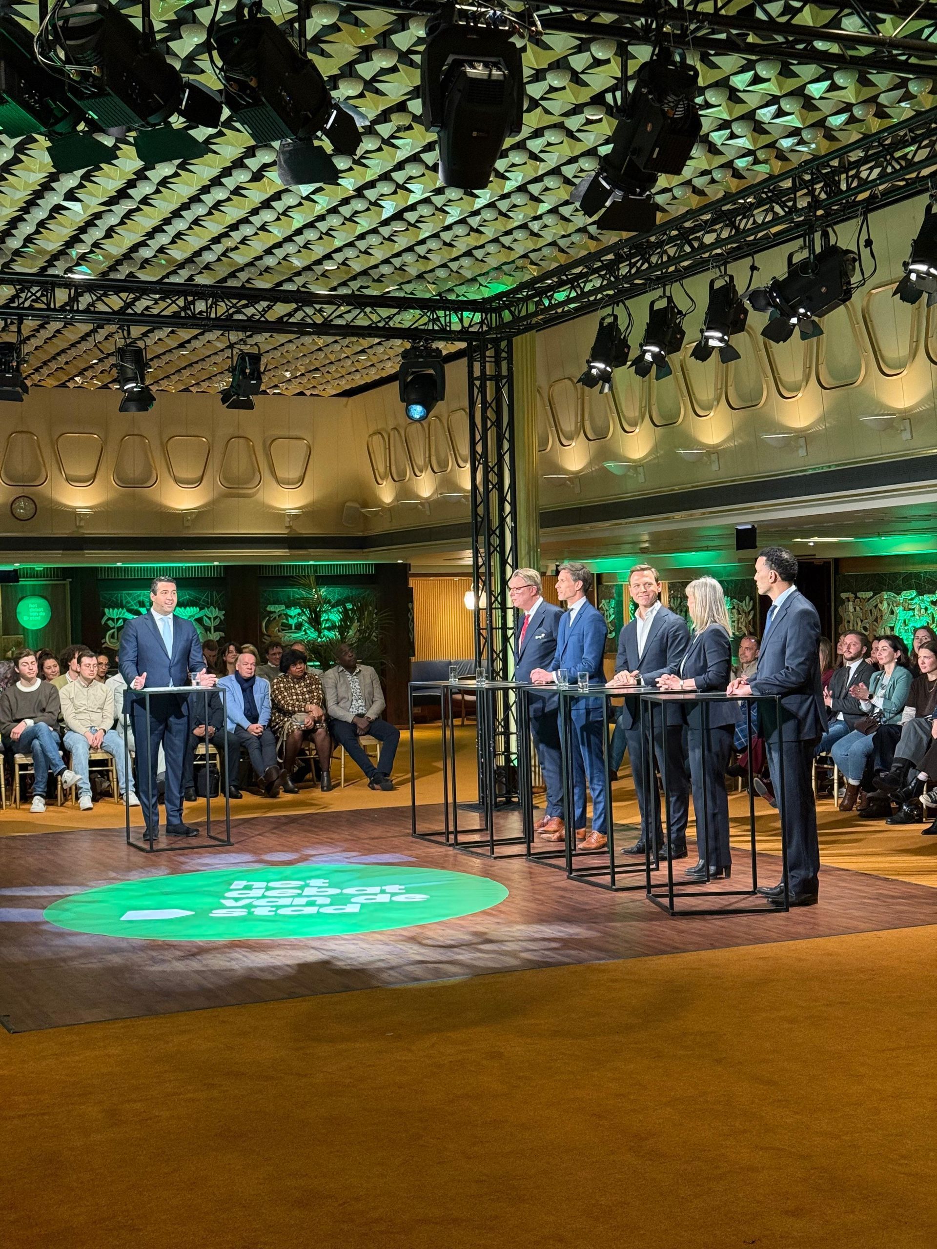 Een panel van mensen staat achter spreekstoelen op een podium met bovenverlichting geeft een presentatie in een vergaderzaal.