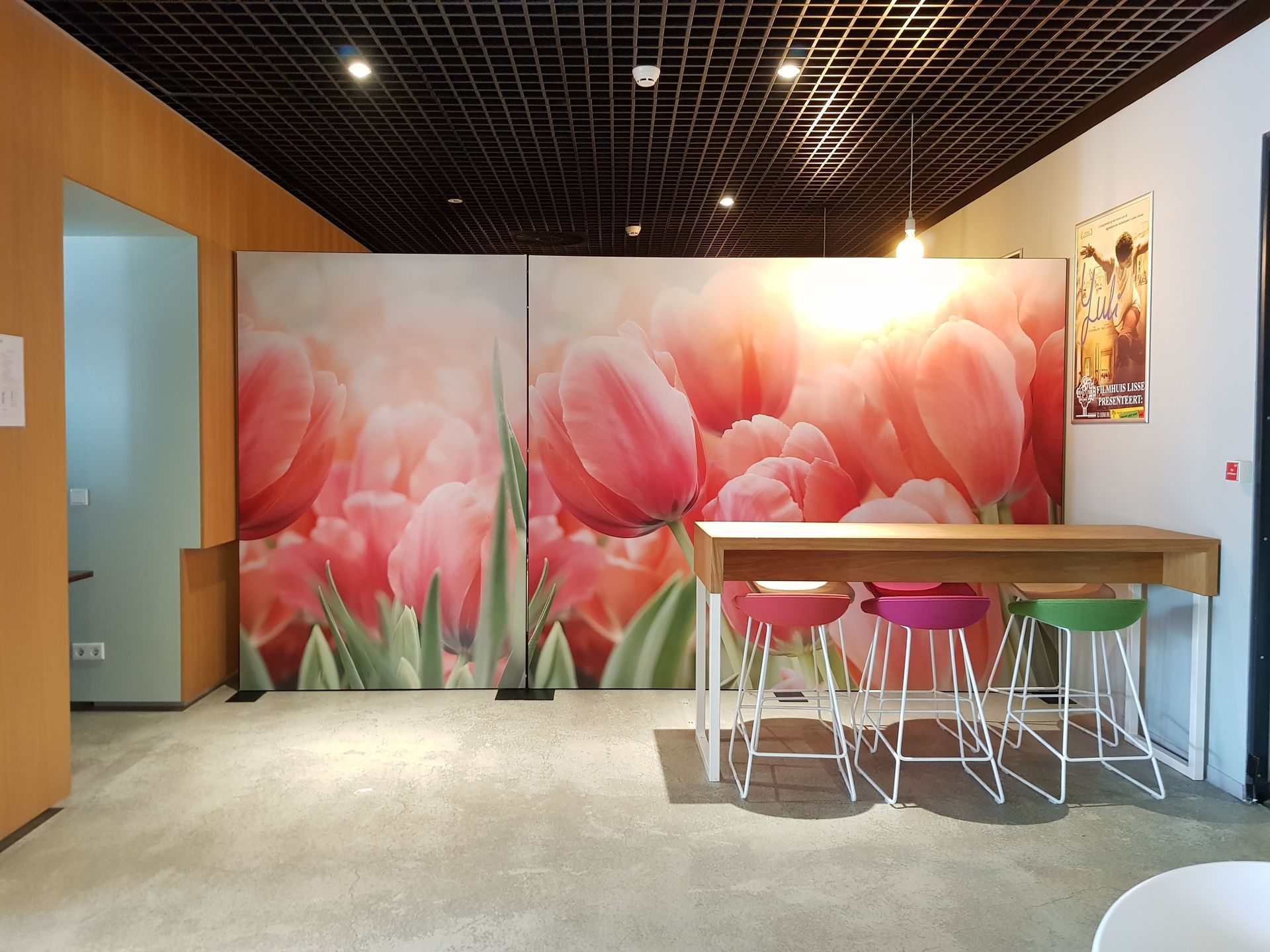 Een hoge houten tafel met rode, roze en groene krukjes voor twee grote panelen met roze tulpen.