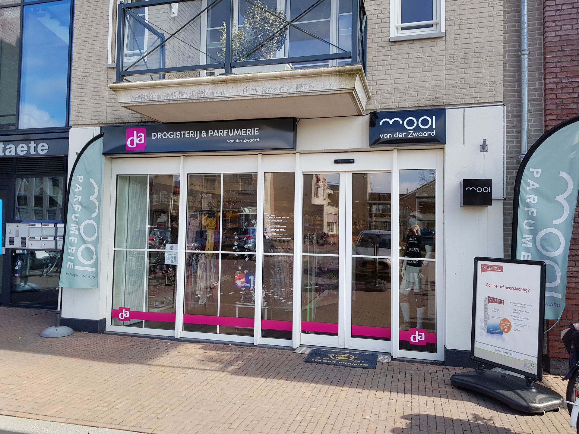 Een winkelpand met een glazen gevel, witte omlijsting en het Mooi-logo, met reclamebanners op een bakstenen stoep.