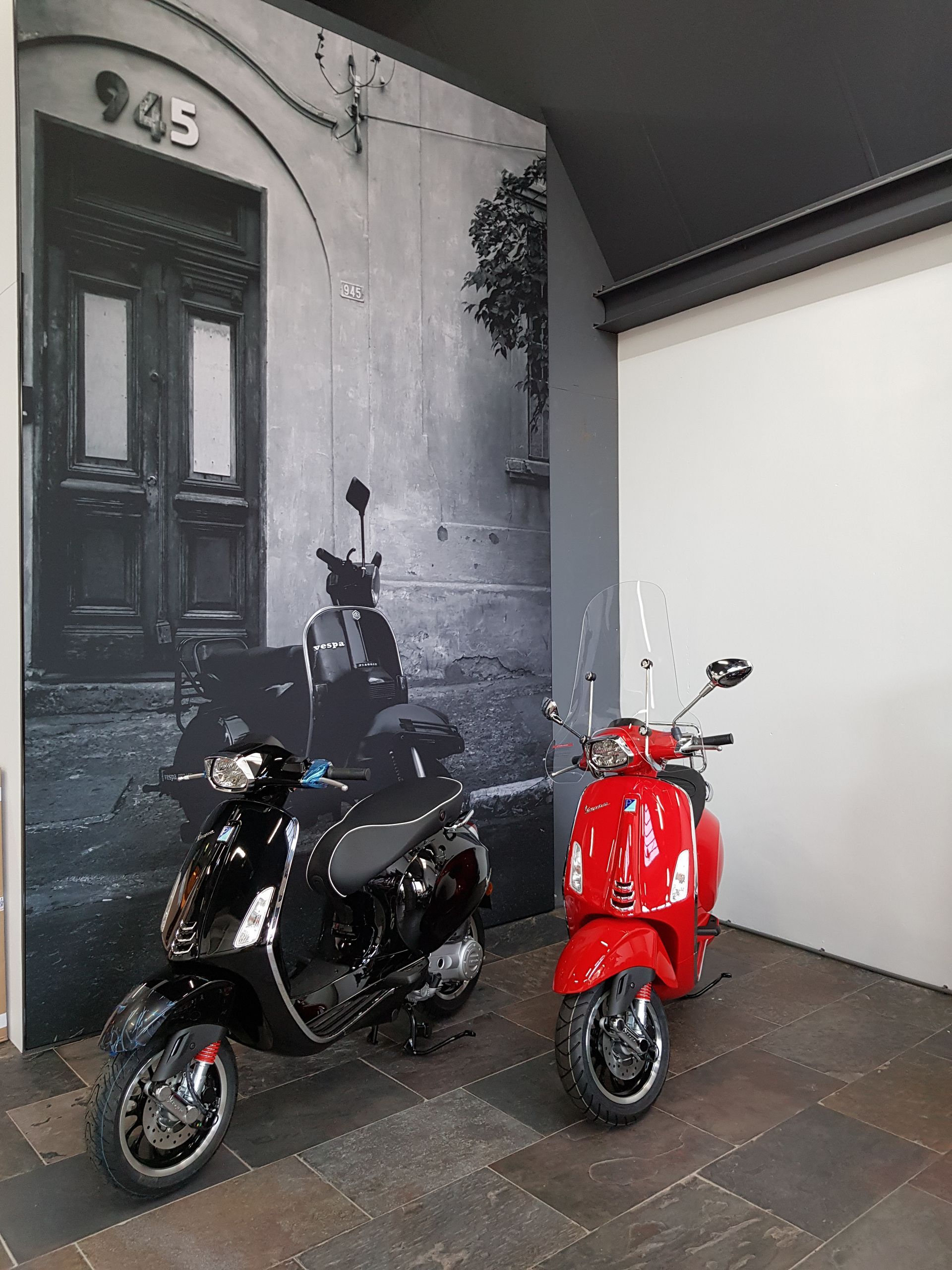 Een zwarte en een rode Vespa-scooter staan ​​geparkeerd voor een muurschildering van een deuropening met het nummer 945.