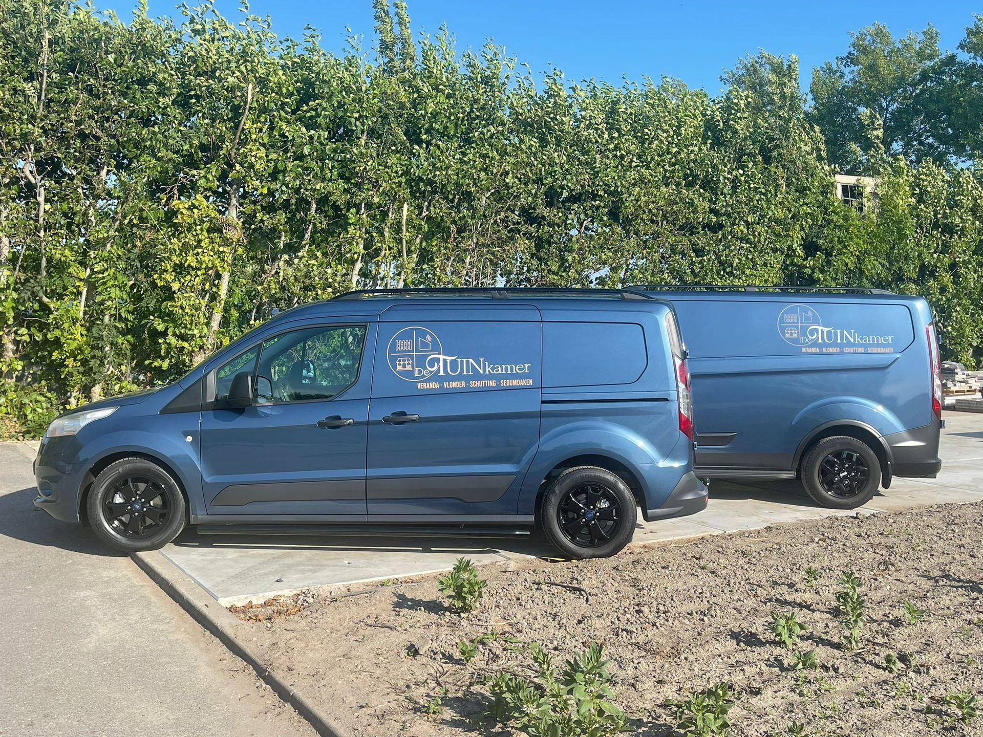 Twee blauwe bedrijfswagens staan ​​naast elkaar geparkeerd op een verharde parkeerplaats voor een dichte groene heg.