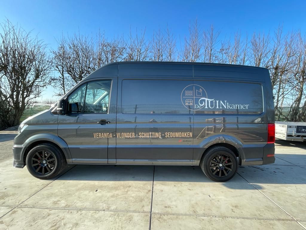 Een donkergrijze bestelbus, met een wit logo en tekst op de zijkant, tegen een zonnige hemel.