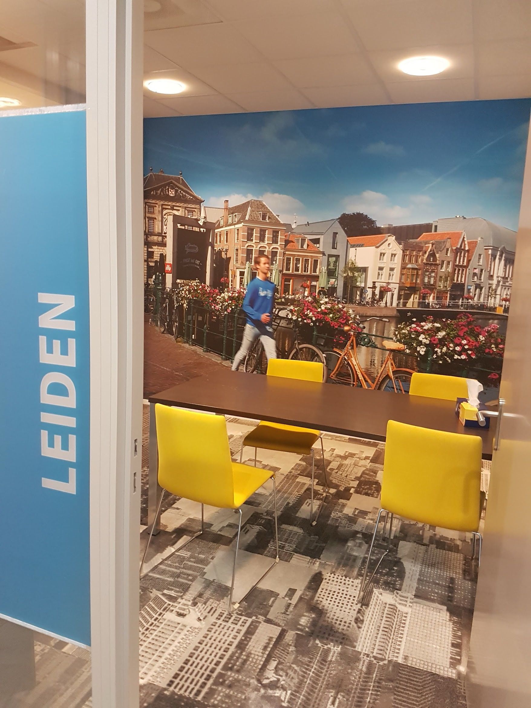 Een vergaderruimte met een muurschildering van een kanaalstad, een donkerbruine tafel en vier felgele stoelen.