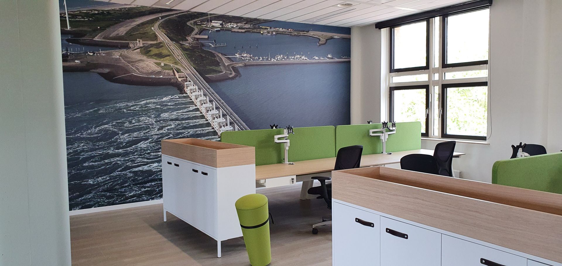 Moderne kantoorruimte met bureaus van licht hout, groene scheidingswanden, witte opbergkasten en een grote muurschildering.