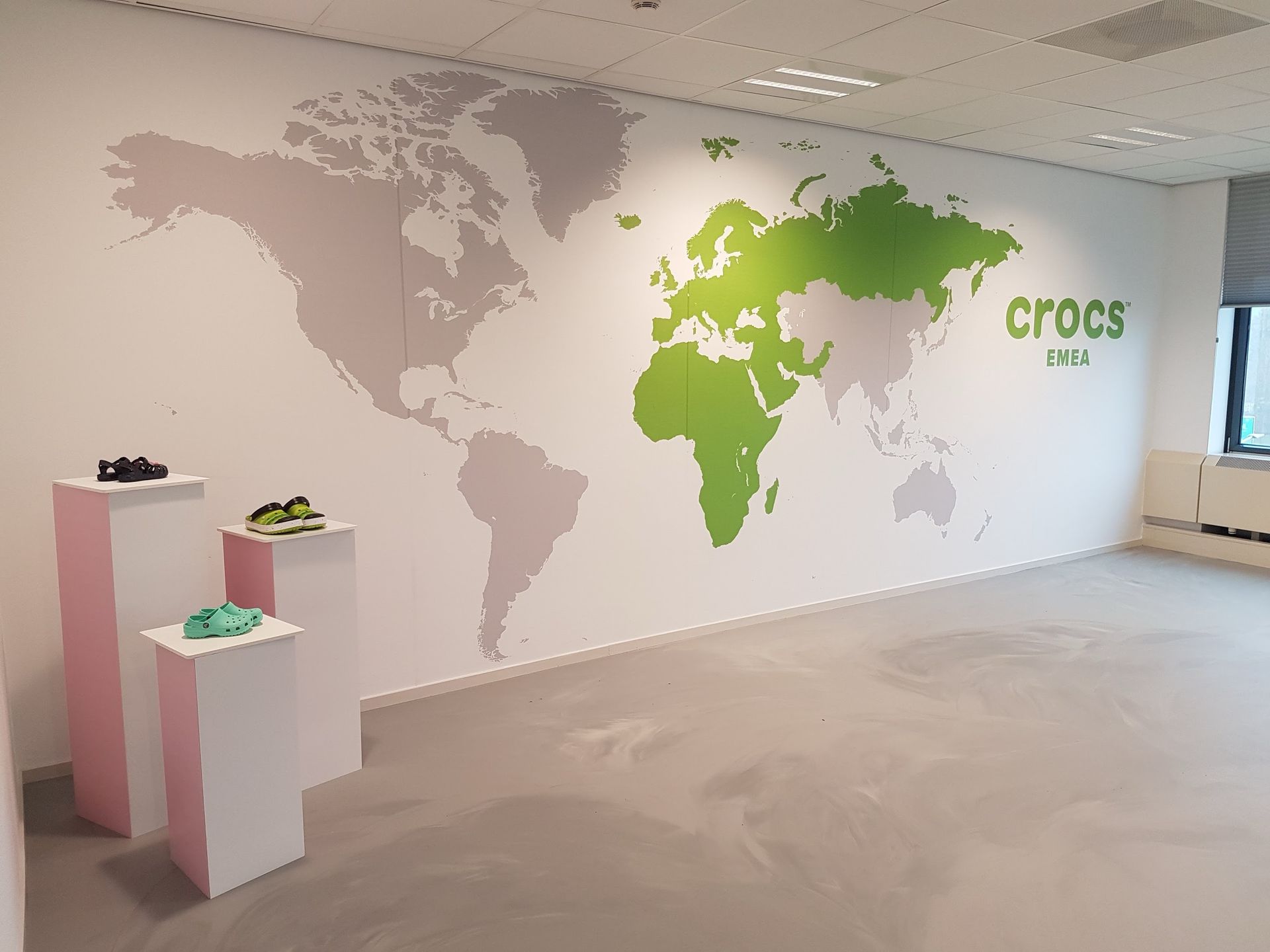 een muur waarop een wereldkaart en het Crocs-logo te zien zijn, en drie sokkels waarop Crocs-schoenen worden tentoongesteld.