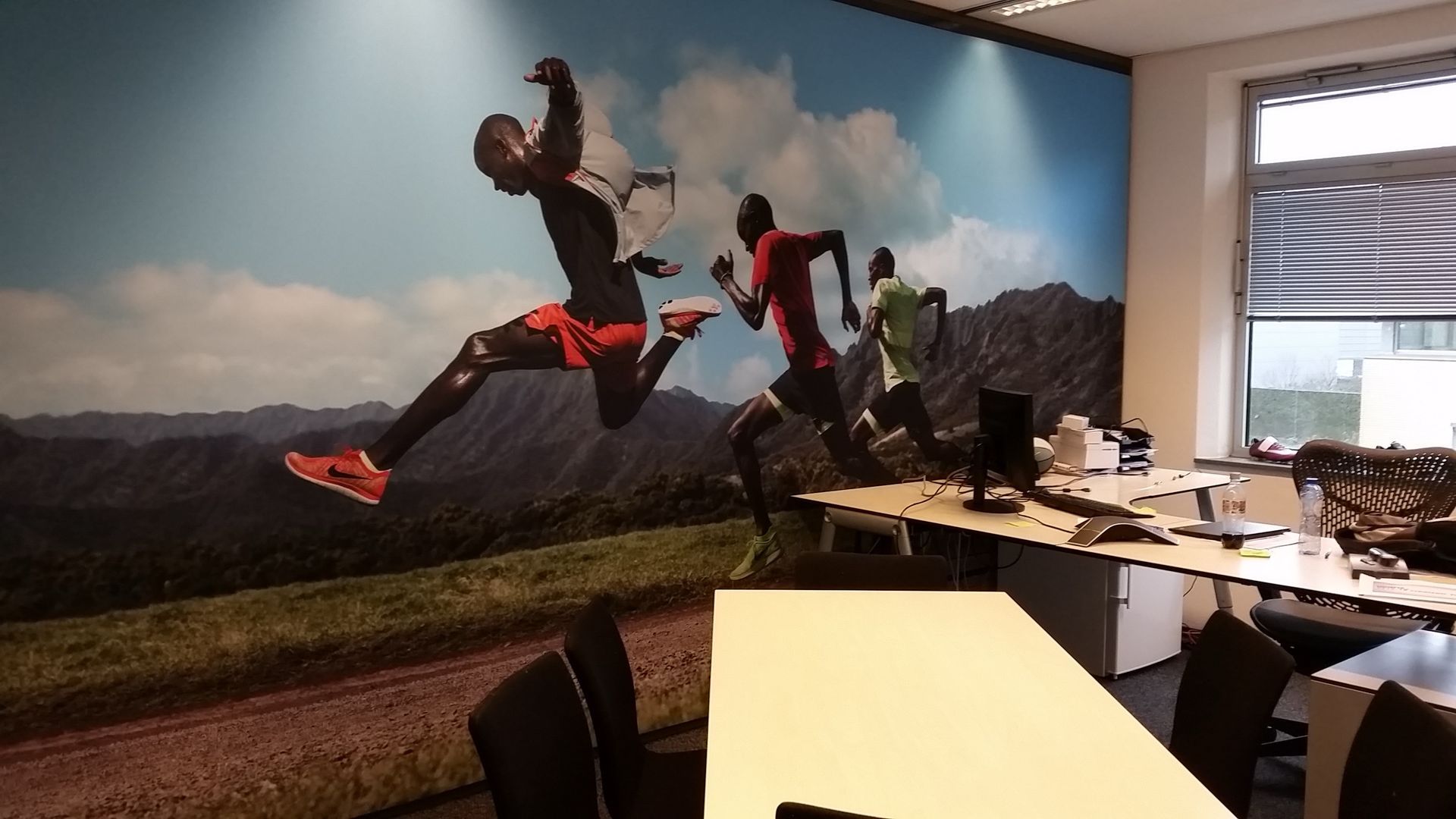 Een wallcover van rennende mensen in beweging tegen in een kantooromgeving met bureaus en stoelen op de voorgrond.