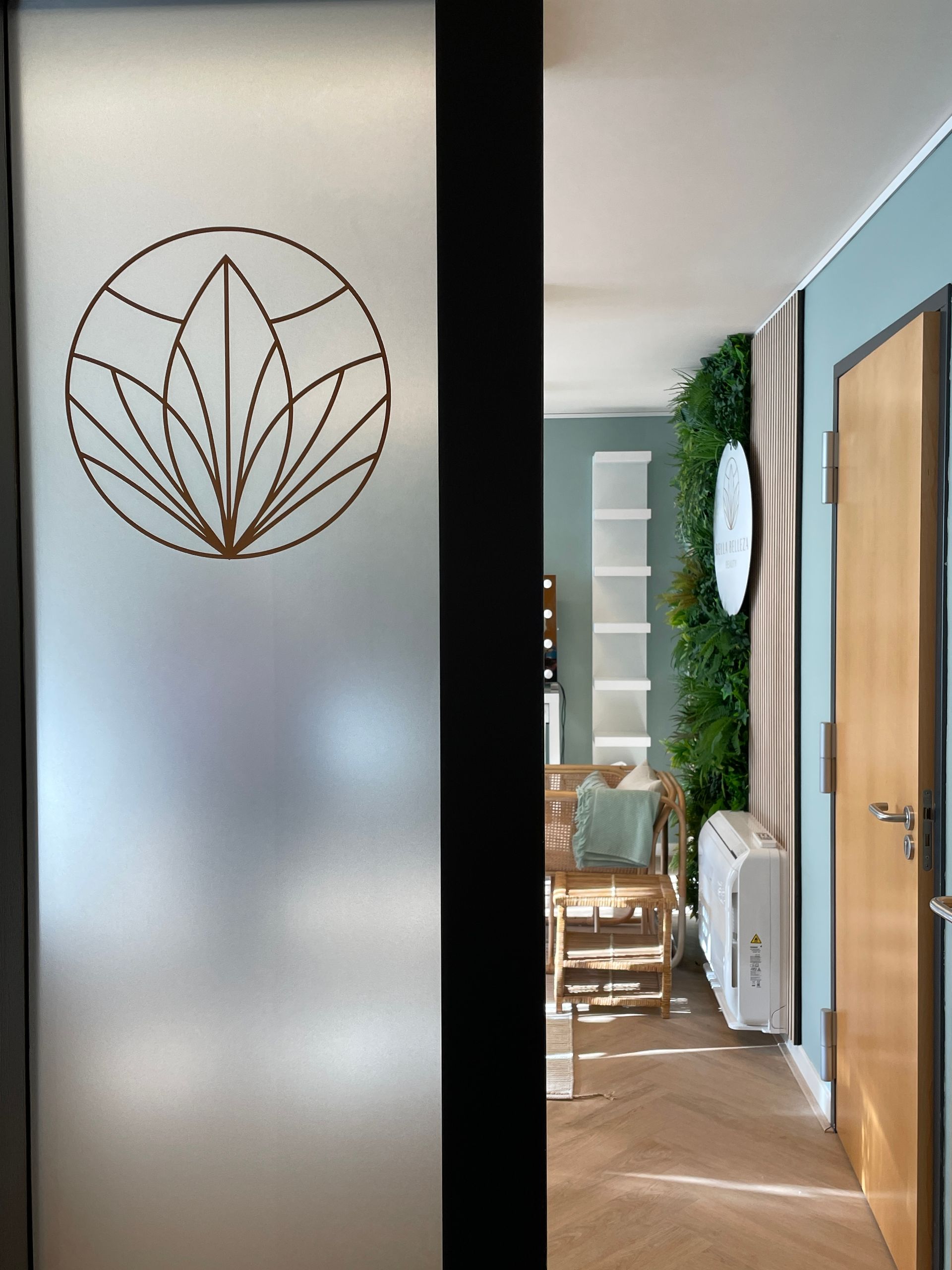 Een deur van matglas met een rond bloemenlogo geeft toegang tot een modern, licht kantoor met planten en houten vloeren.