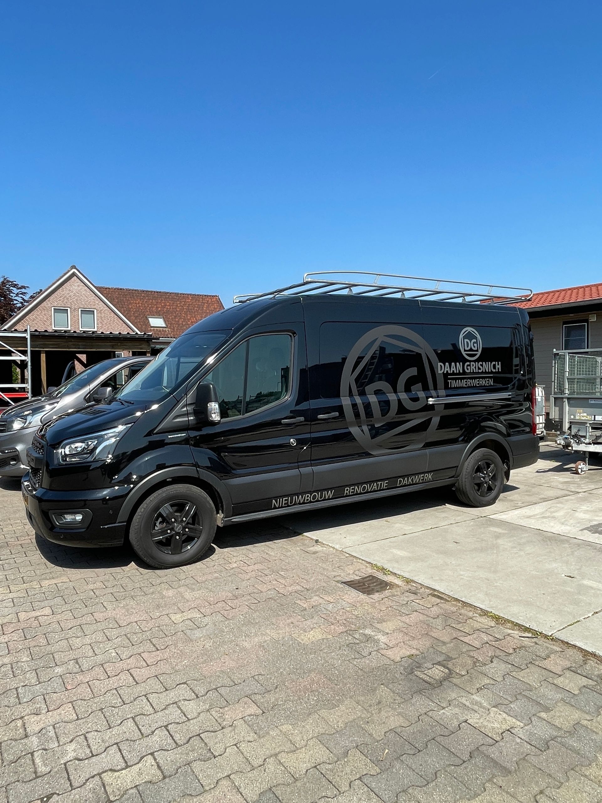 Een zwarte bestelbus met imperiaal en logo staat geparkeerd op een geplaveide parkeerplaats tegen een helderblauwe hemel.