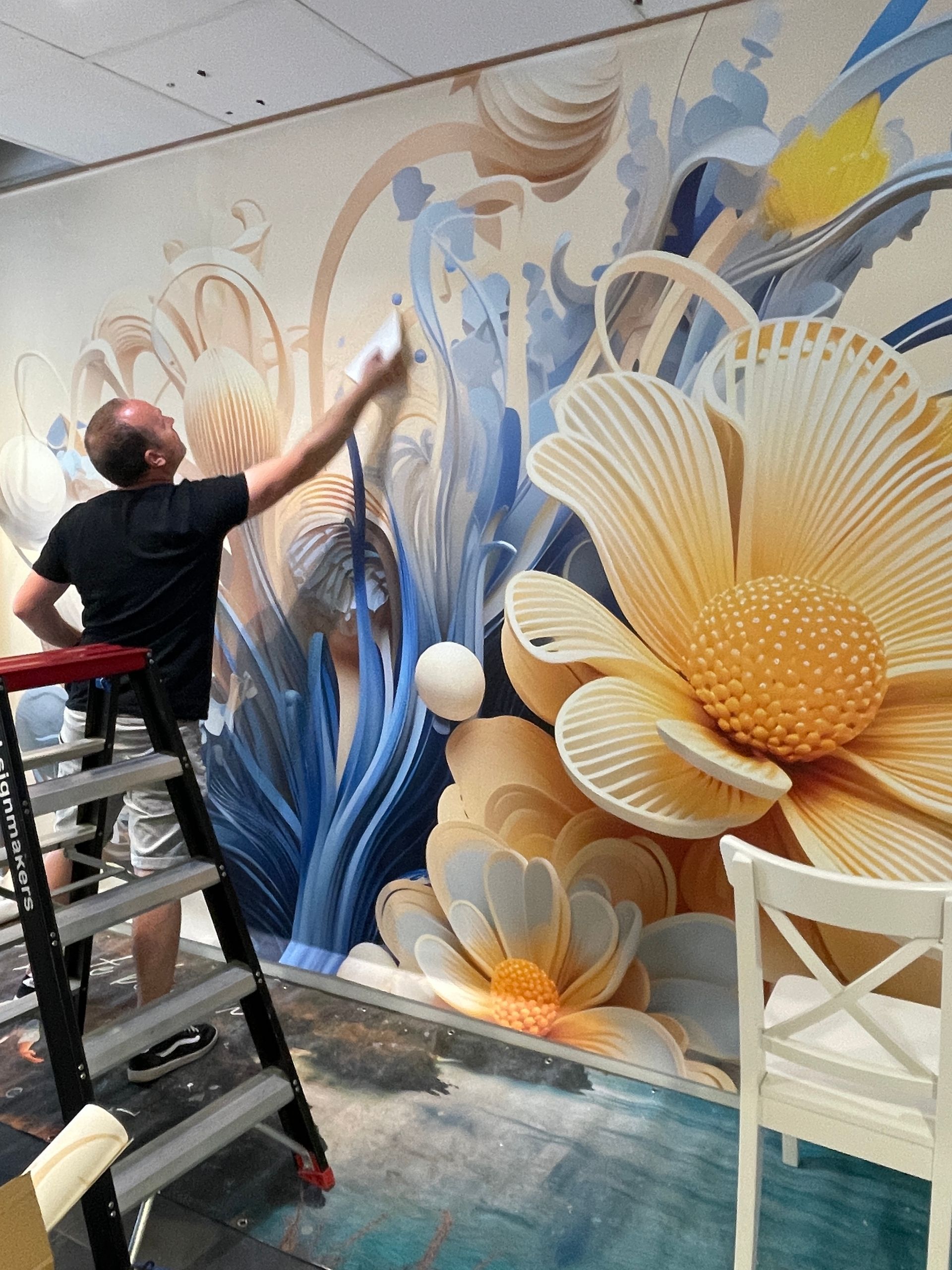 Iemand staat op een ladder en schildert een grote, gedetailleerde muurschildering met blauwe versieringen en gele bloemen.