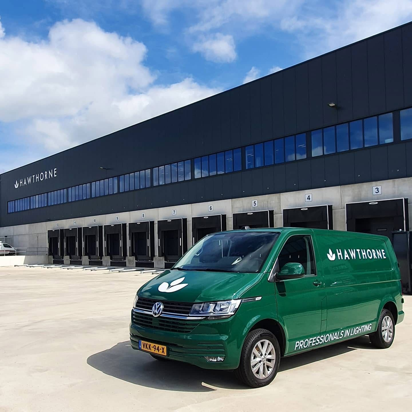 Een groene bestelbus staat geparkeerd voor een modern, donkergekleurd magazijngebouw met meerdere laadperrons.