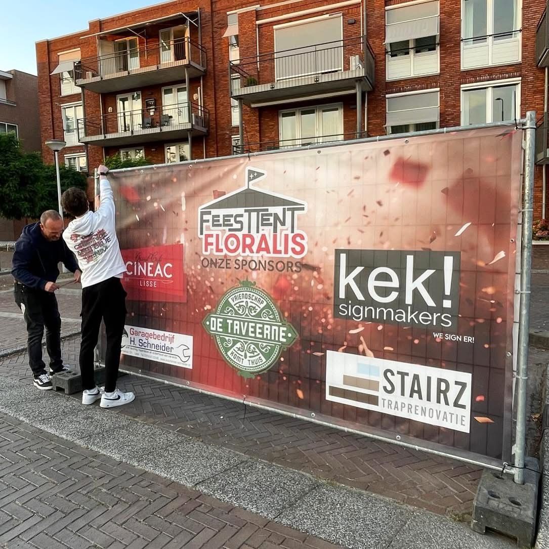 Twee personen bevestigen een spandoek met de namen van de sponsors van het evenement aan een tijdelijk metalen hek.