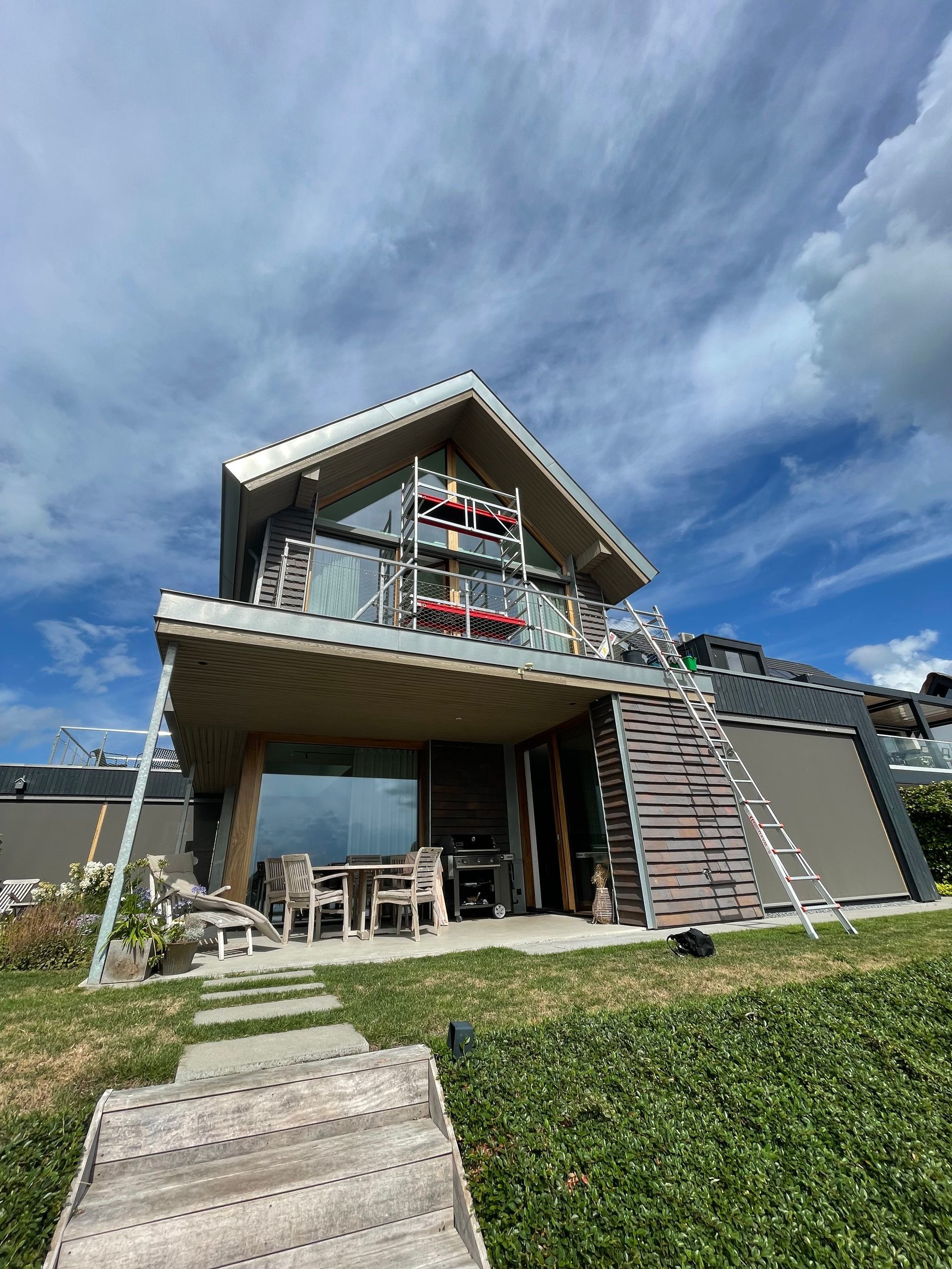 Een woning met twee verdiepingen, een balkon en grote ramen, met een schuin dak onder een heldere, bewolkte blauwe hemel.