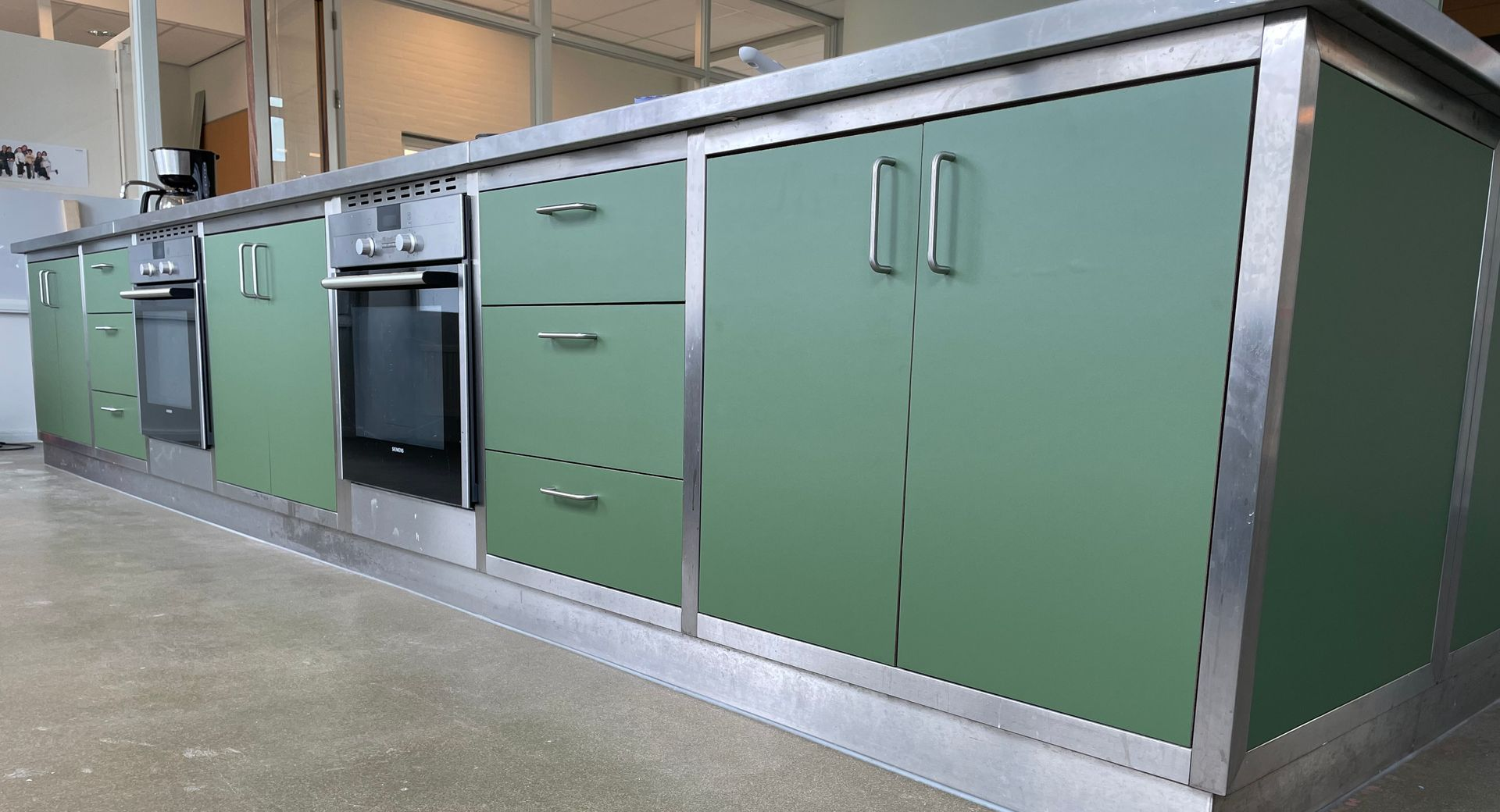 it's all about the details Modern keukeneiland met matgroene kasten, roestvrijstalen afwerking en twee ingebouwde ovens op een betonnen vloer.