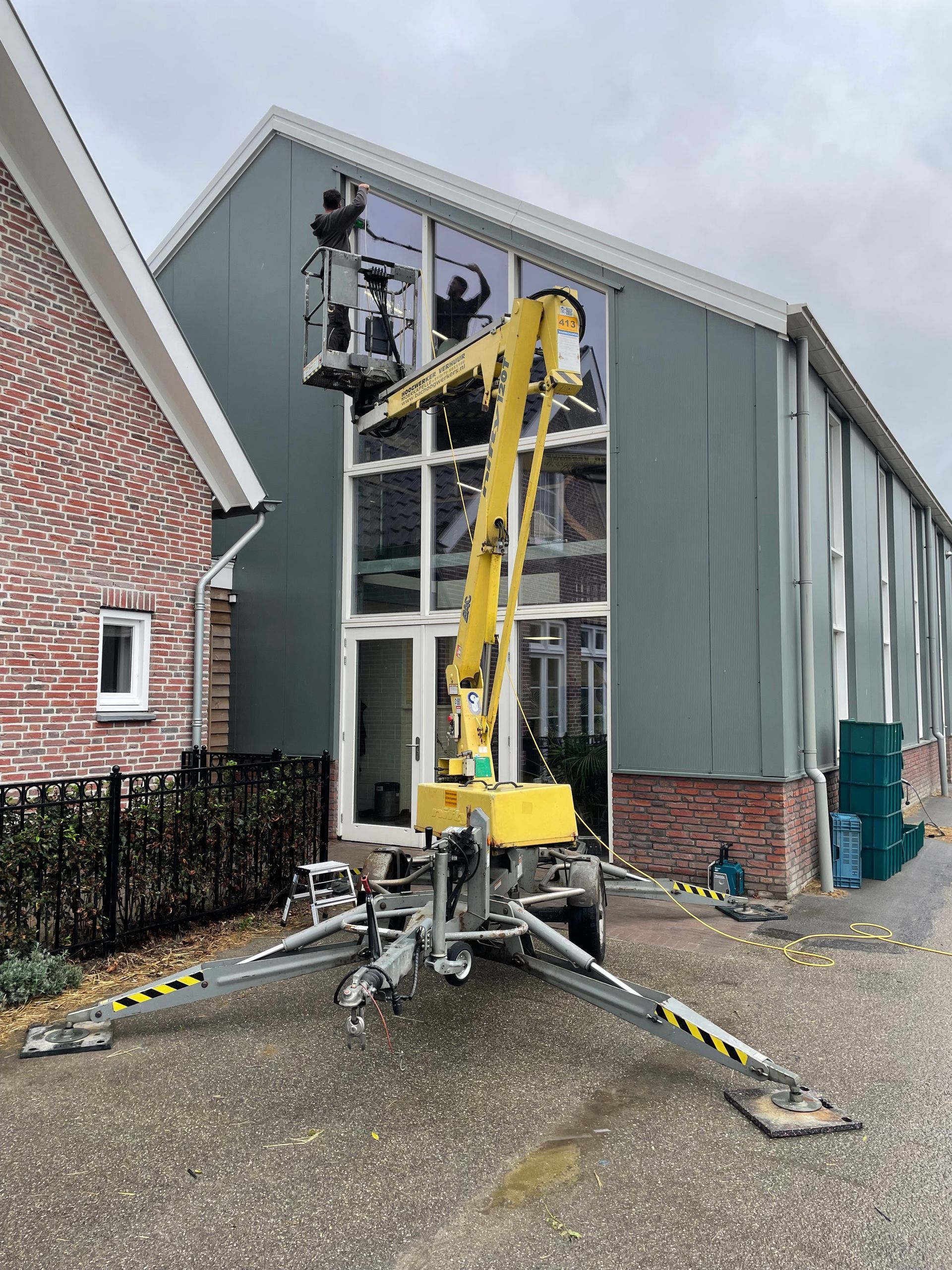 Een geel hoogwerkplatform strekt zich uit naar de hoge, met glas beklede gevel van een gebouw met grijze gevelbekleding.