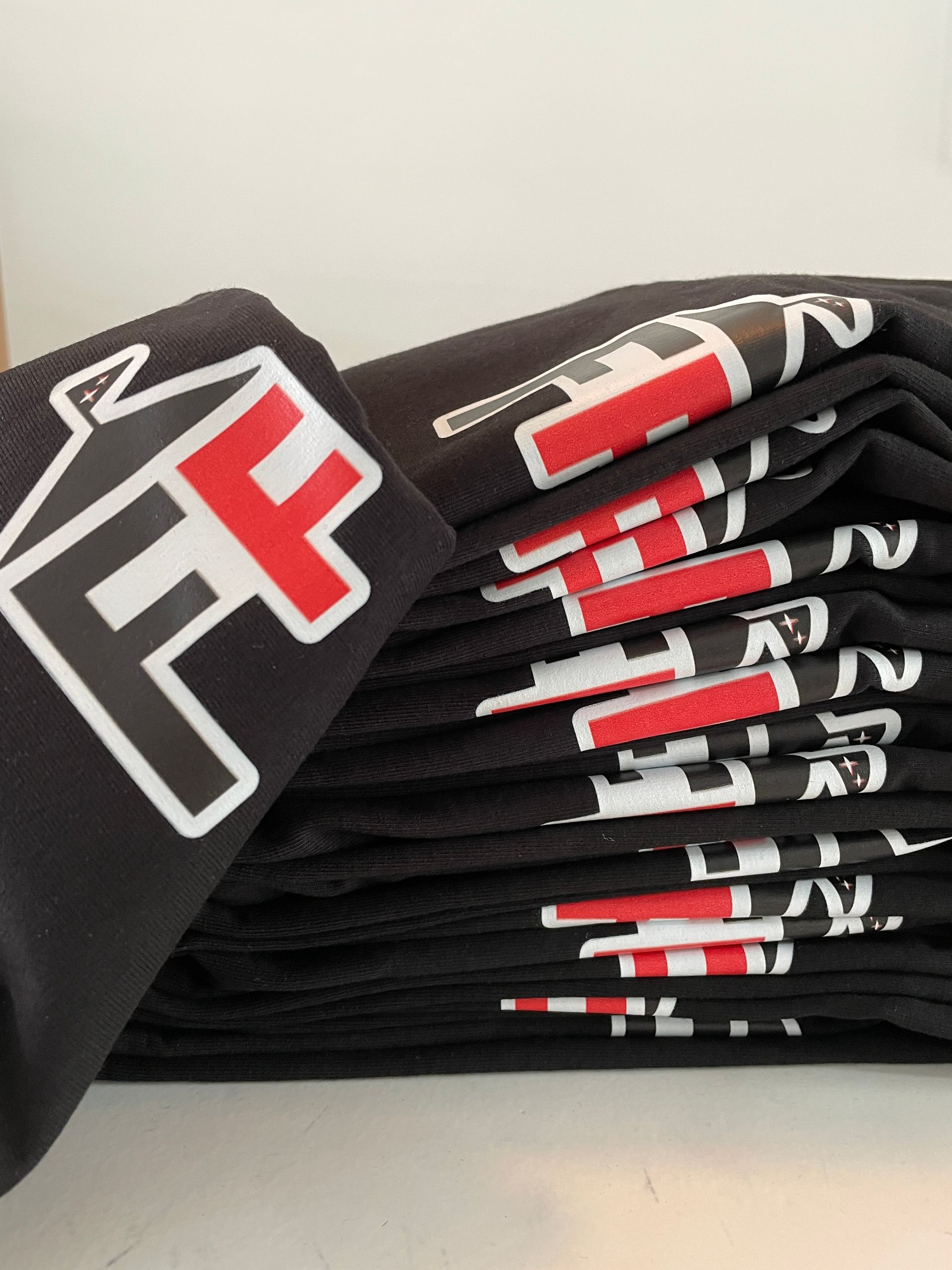 Een stapel zwarte T-shirts, elk voorzien van een logo met een witte omtrek waarin de letters 