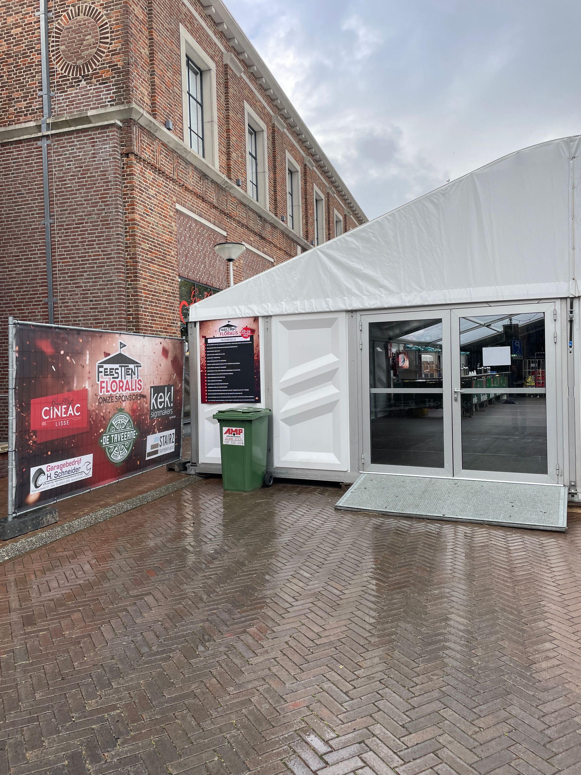 Naast een bakstenen gebouw staat een witte evenemententent, met een groene afvalbak en reclameborden op een geplaveid plein.