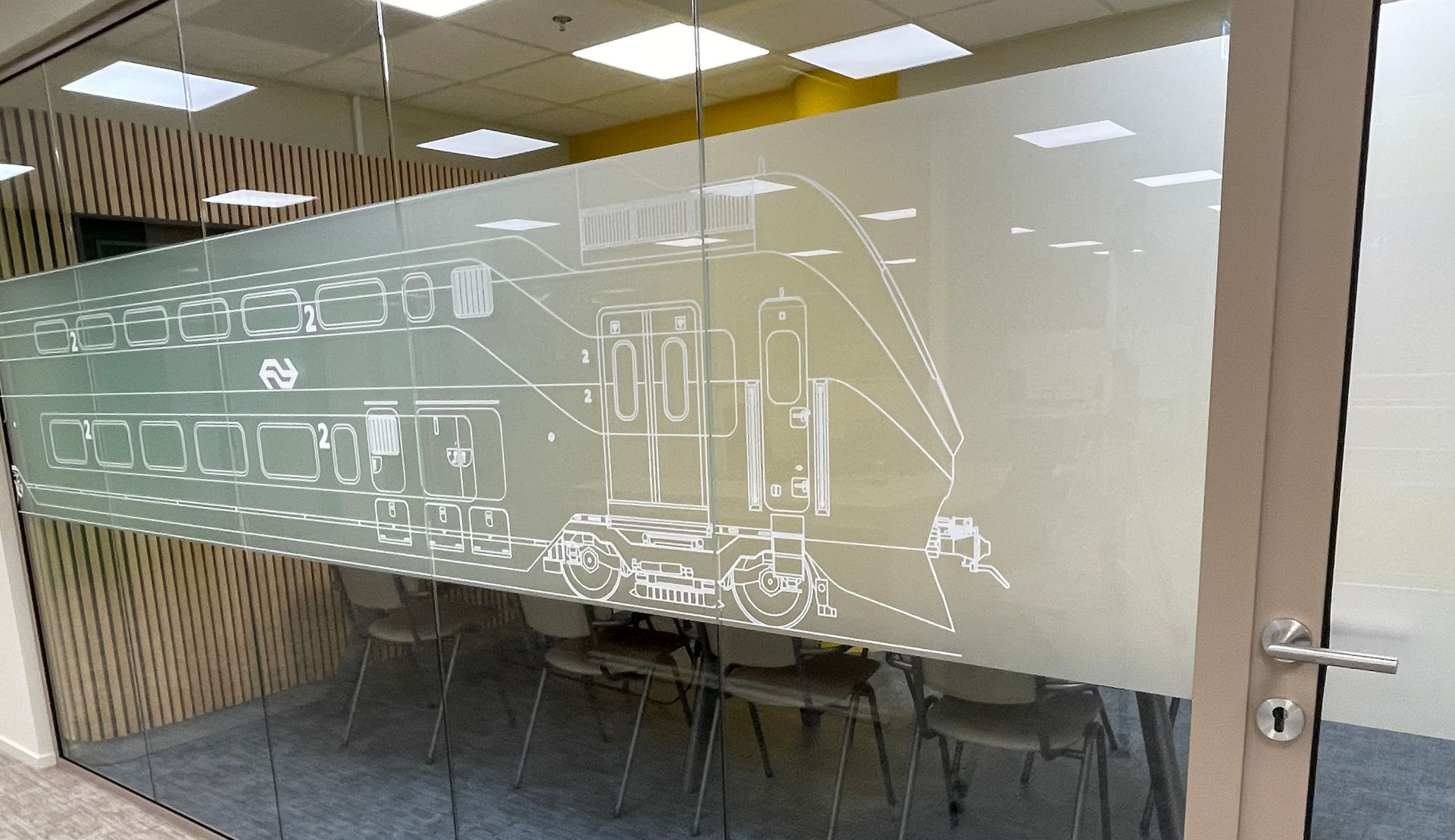 it's all about the details Op een kantoordeur van matglas is een witte lijntekening te zien van een dubbeldekkertrein, met stoelen binnenin zichtbaar.