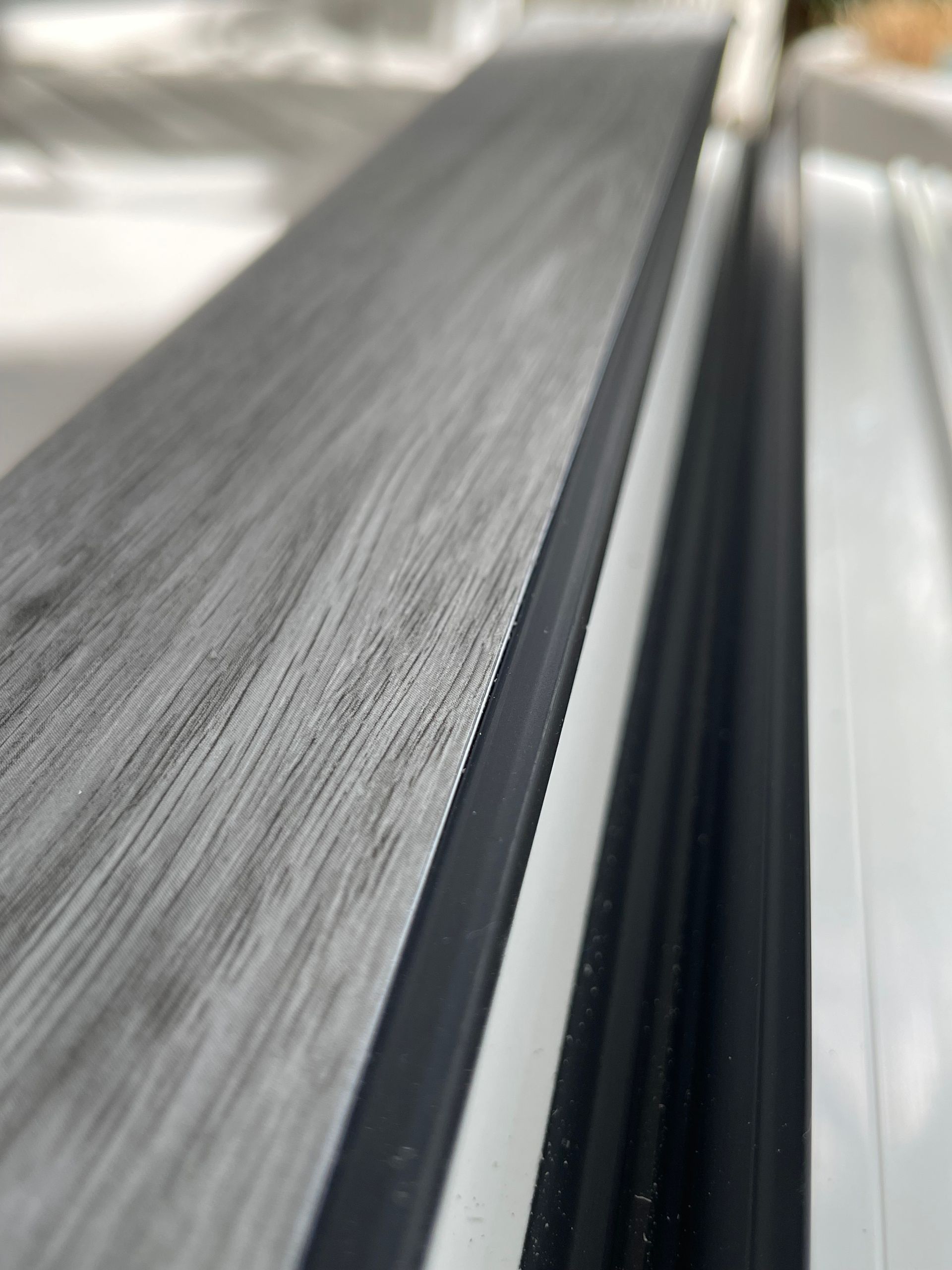 Een close-up, schuin genomen, toont een grijs houtnerfoppervlak naast zwarte rubberen tochtstrips en een witte frame-rand.