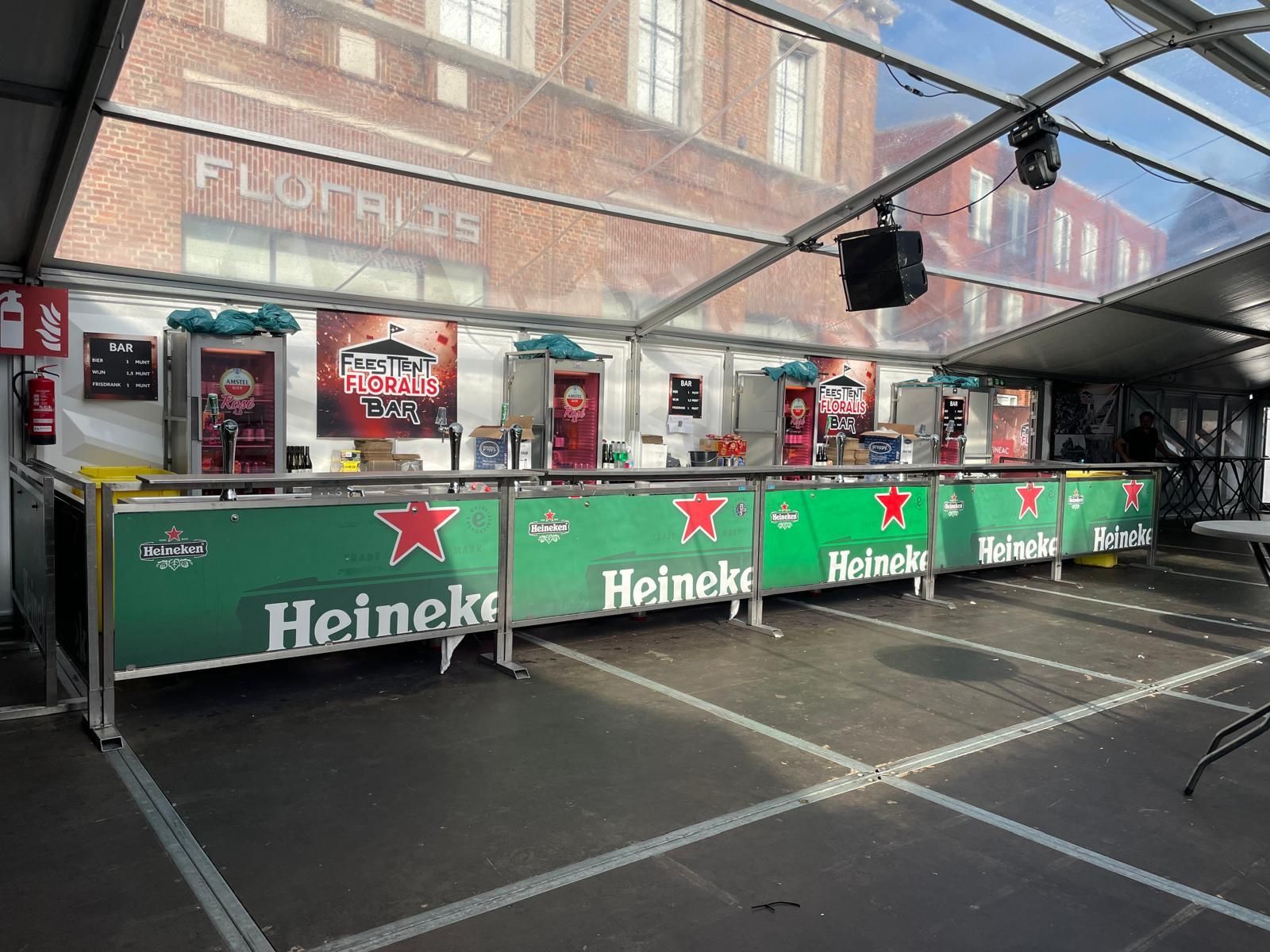 Een lange bar met Heineken-logo, opgesteld in een tent met een transparant dak, met meerdere bierkoelers.