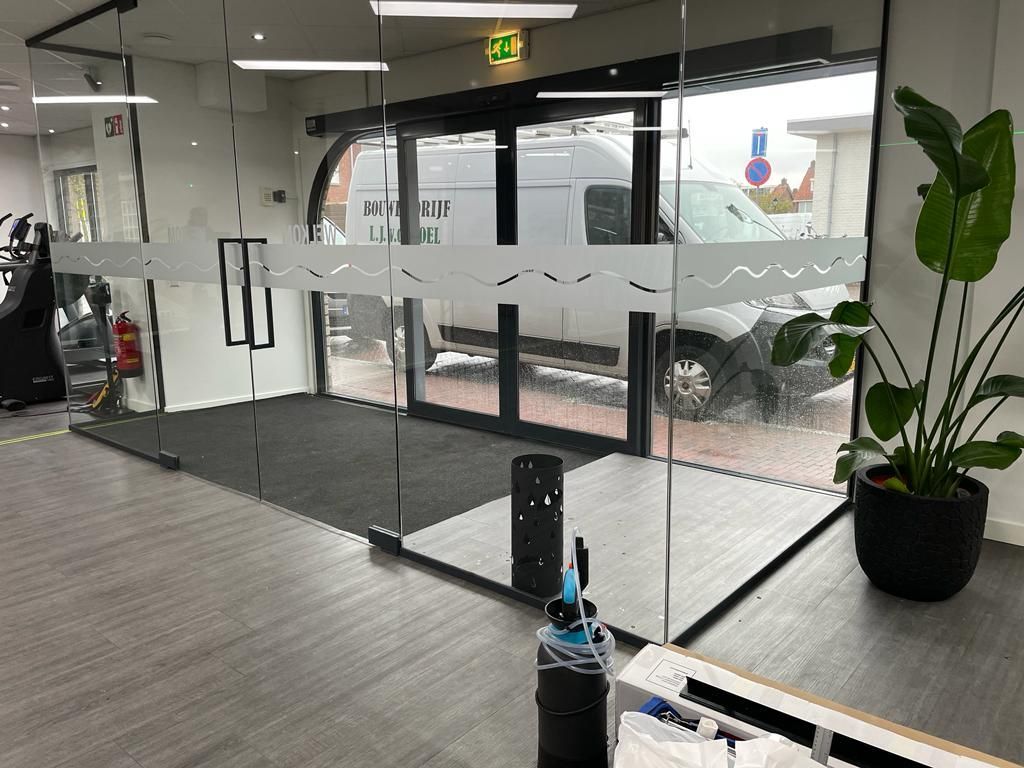 Een glazen entree met schuifdeuren in een sportschool, een potplant en een witte bestelwagen die buiten zichtbaar is.