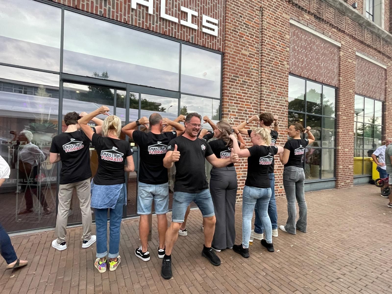 Een groep mensen, allemaal in dezelfde zwarte T-shirts, spant hun armen voor een bakstenen gebouw met grote glazen ramen.