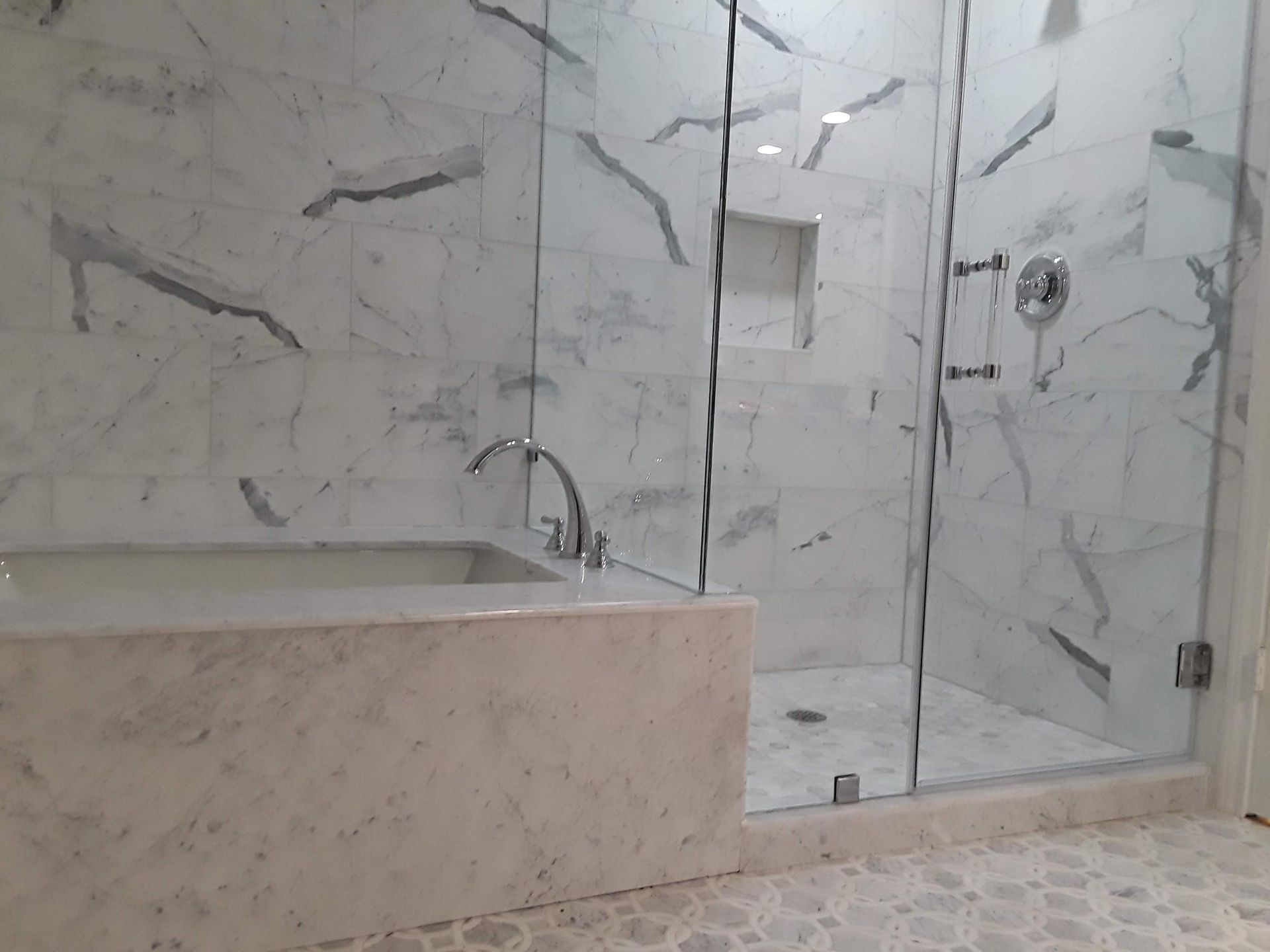 Luxurious bathroom with marble walls and glass shower. Bathtub, showerhead, and a small recessed shelf.