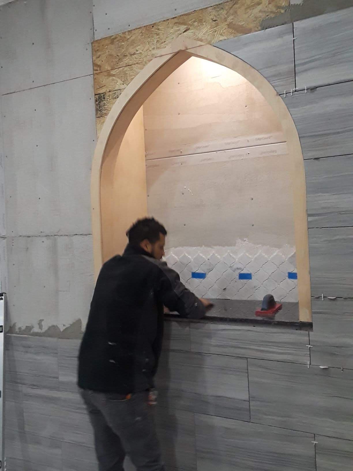 Person tiling around an arched alcove. Walls are gray tile and drywall.